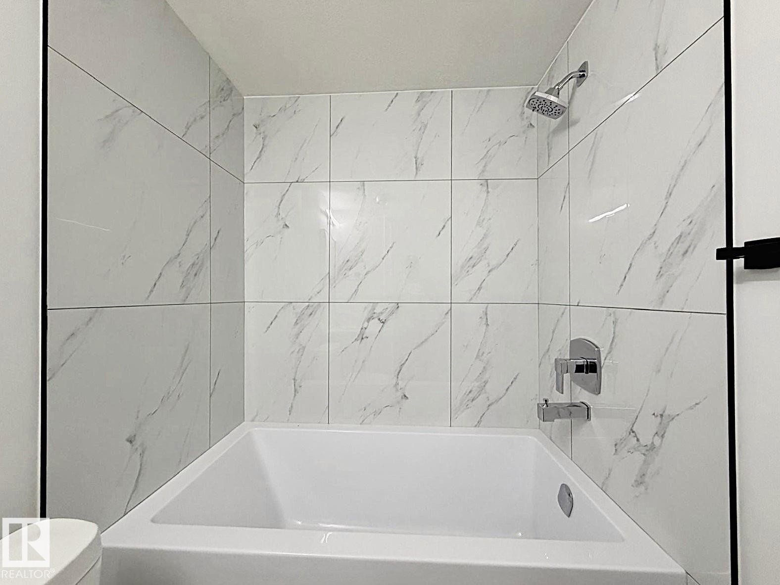 Bathtub with a shower system featuring a handheld showerhead, chrome fixtures, and white tile surround with subtle gray veining - 1312 35 Street Nw, Edmonton, AB