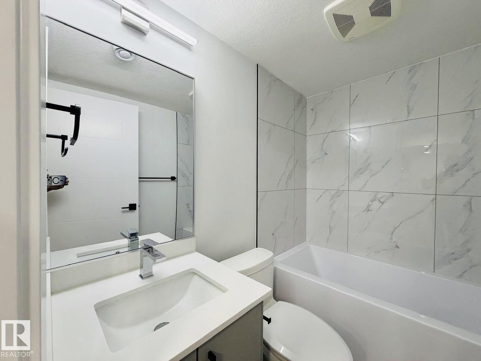 Modern bathroom featuring a rectangular vanity sink with a chrome faucet, a full-width wall mirror, and a white bathtub surrounded by white marble-look tile - 1312 35 Street Nw, Edmonton, AB