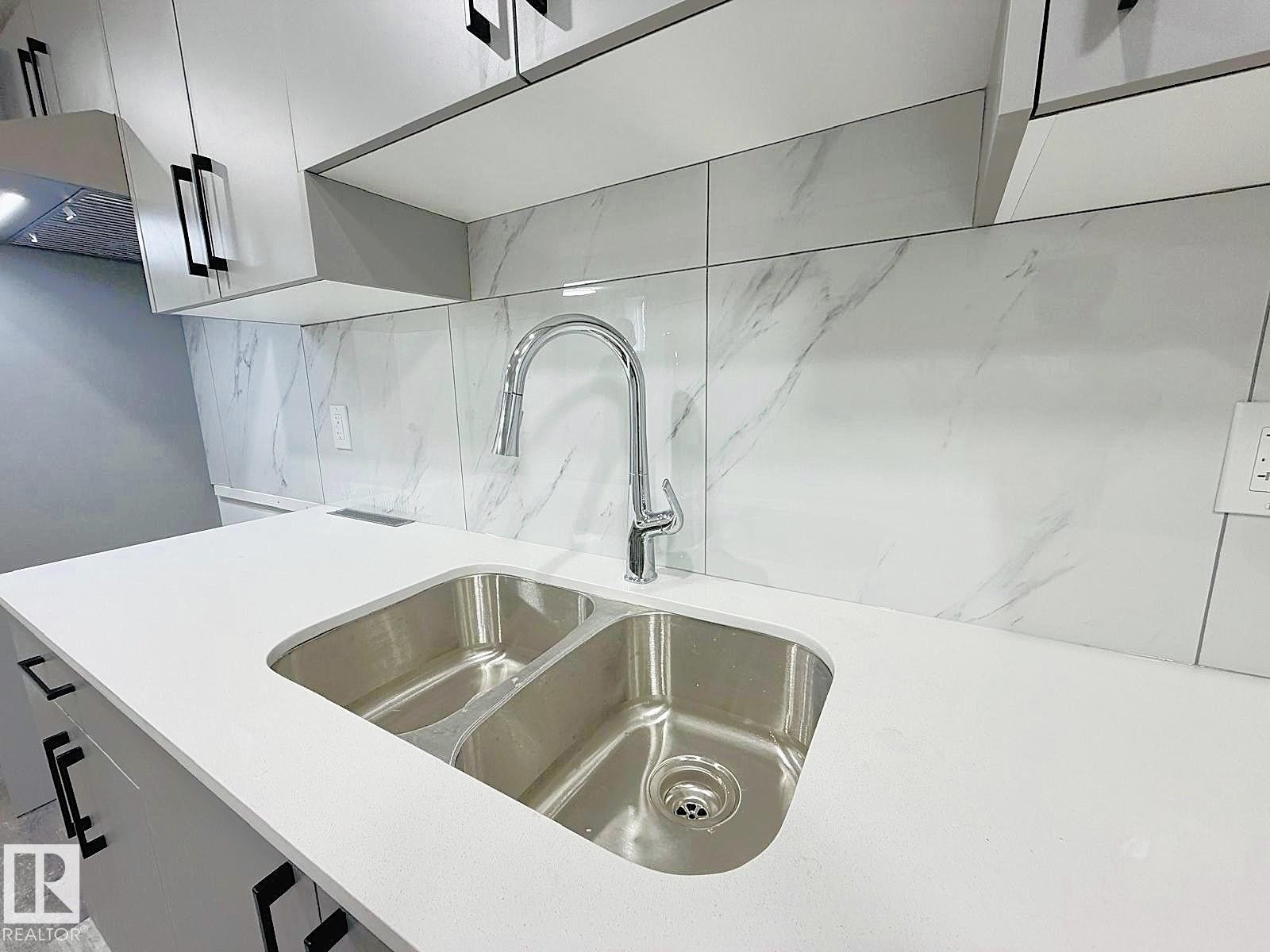 Modern kitchen featuring a double-basin stainless steel sink, pull-down faucet, white countertop, white and gray patterned tile backsplash, and light-toned cabinetry with black hardware - 1312 35 Street Nw, Edmonton, AB