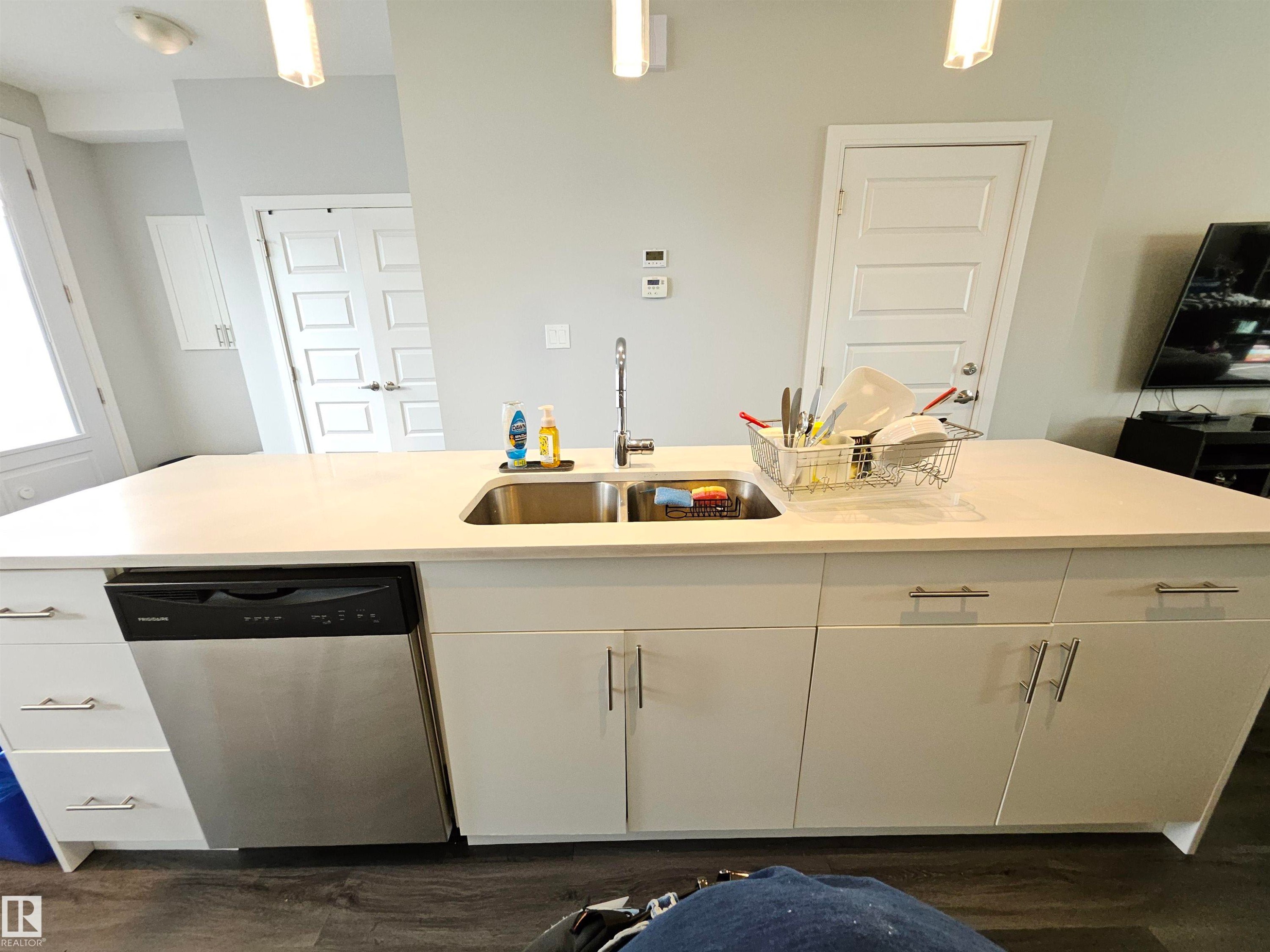 Kitchen island featuring a stainless steel double-basin sink, integrated dishwasher, ample cabinetry, and white countertops - 15212 98 Avenue Nw, Edmonton, AB - Indoor Photo Showing Kitchen With Double Sink