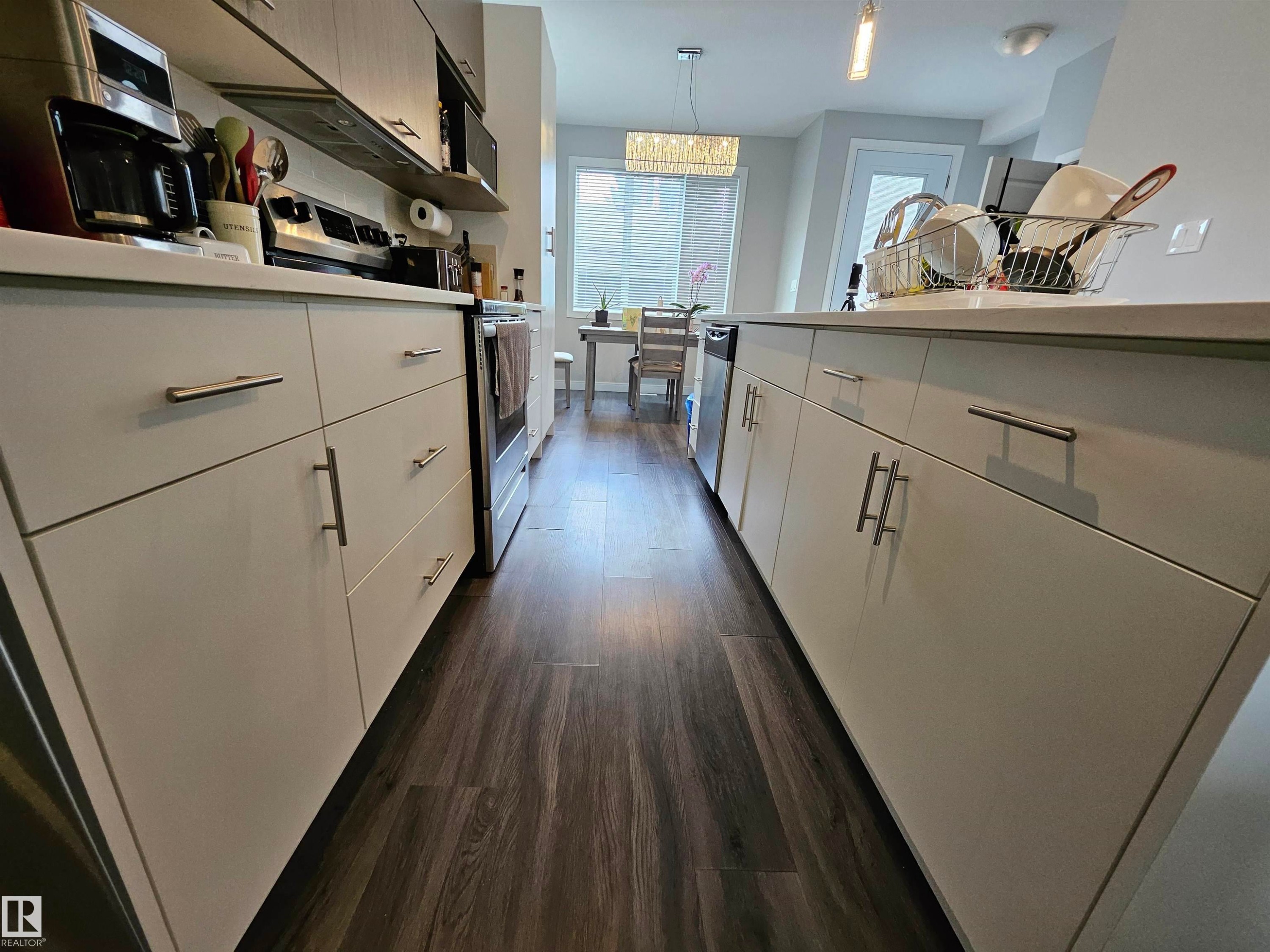 Galley-style kitchen featuring white cabinetry with brushed metal hardware, stainless steel appliances including a wall-mounted oven, dark wood-finish flooring, and ample counter space with integrated storage throughout - 15212 98 Avenue Nw, Edmonton, AB - Indoor Photo Showing Kitchen