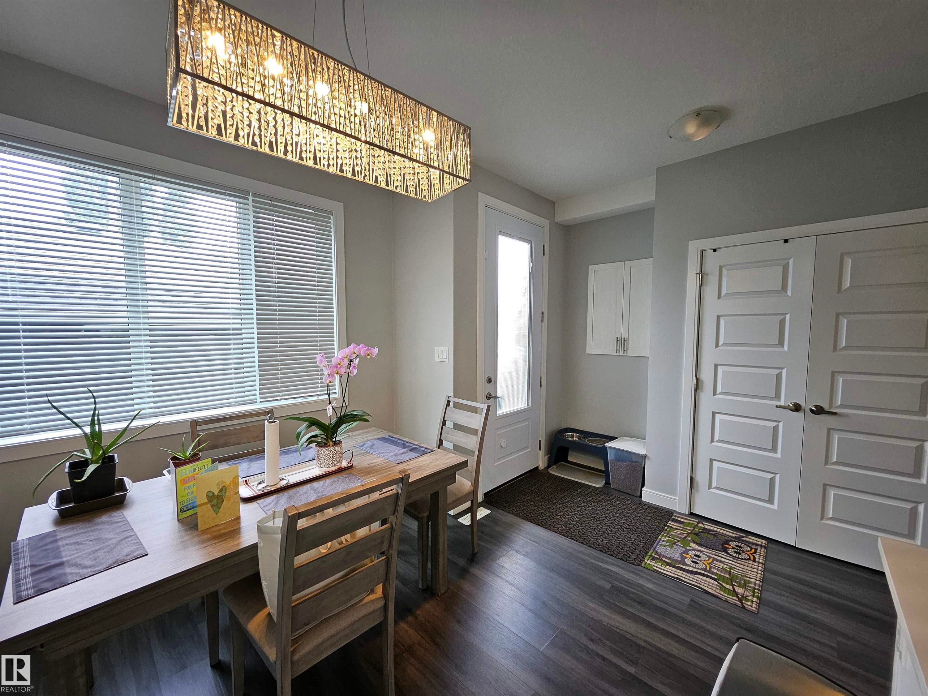 Bright entryway and home office featuring hardwood flooring, a rectangular desk with integrated storage drawers, geometric patterned pendant lighting, white panel closet doors, and natural light from expansive front-facing windows with horizontal blinds - 15212 98 Avenue Nw, Edmonton, AB - Indoor Photo Showing Dining Room