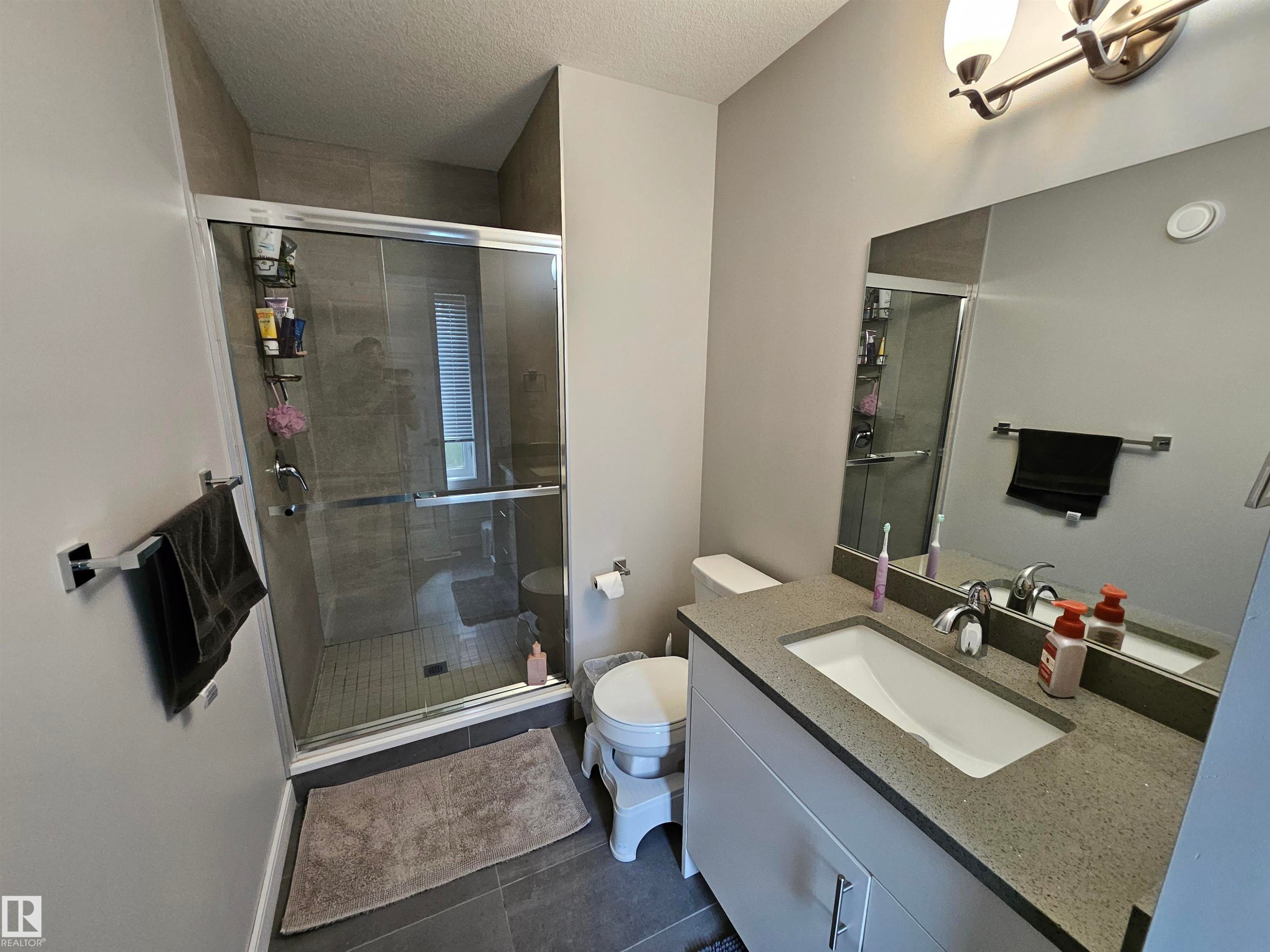 Modern bathroom featuring a frameless glass-enclosed shower with tile surround, dual vanity with quartz countertop and rectangular undermount sink, large mirror with integrated lighting, and neutral tile flooring throughout - 15212 98 Avenue Nw, Edmonton, AB - Indoor Photo Showing Bathroom