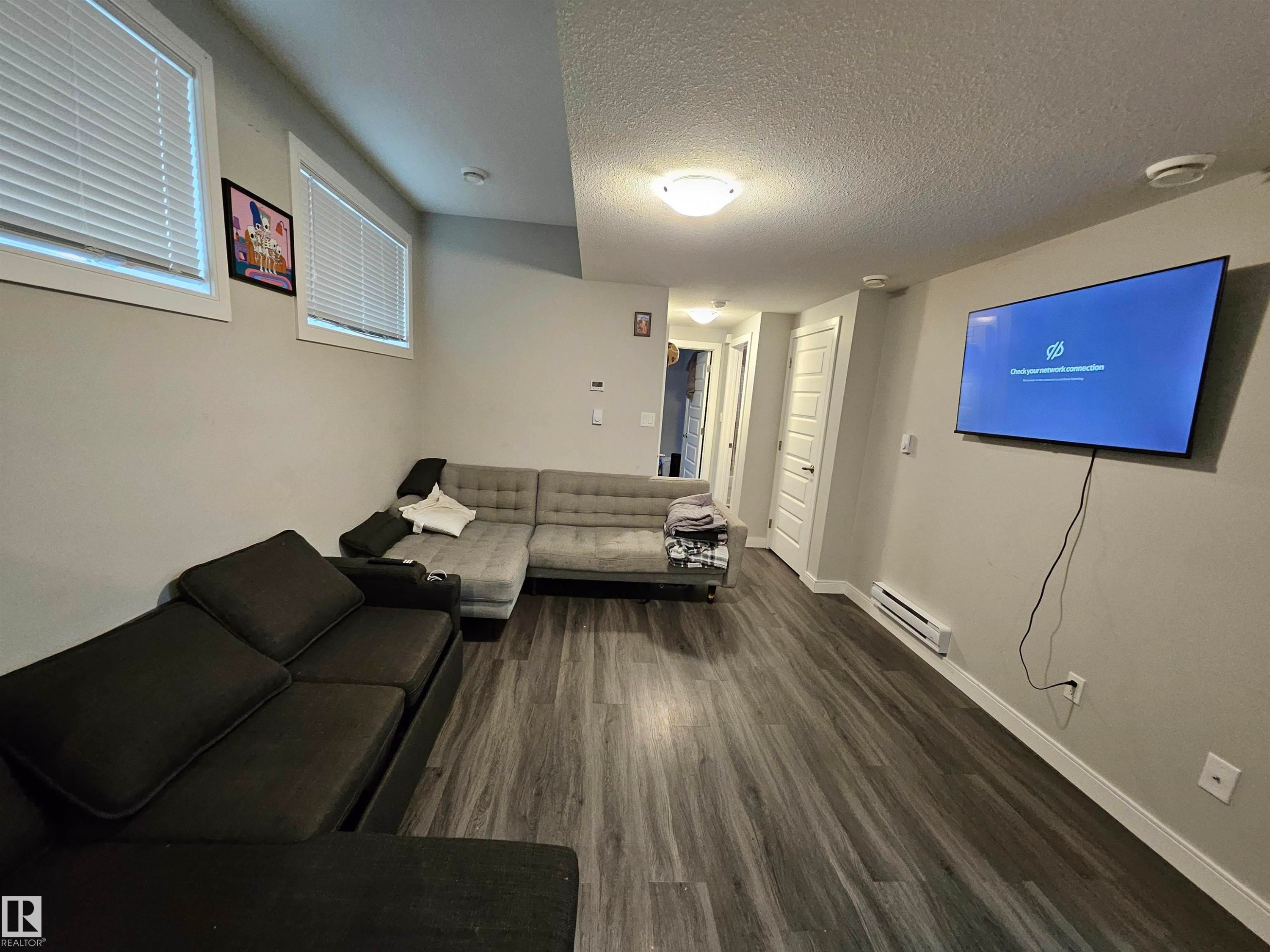 Wood-finish flooring, two windows with blinds, wall-mounted television, and a built-in baseboard heater - 15212 98 Avenue Nw, Edmonton, AB - Indoor