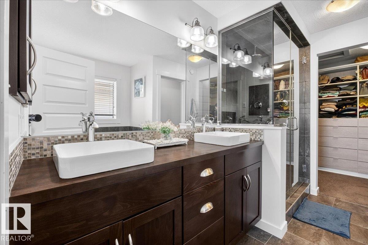 Spa-inspired primary bathroom featuring dual vessel sinks with wood-finish vanity cabinetry, quartz countertops with mosaic tile backsplash, chrome fixtures, and large-format mirror with integrated lighting - 3936 Claxton Loop Sw, Edmonton, AB - Indoor Photo Showing Bathroom