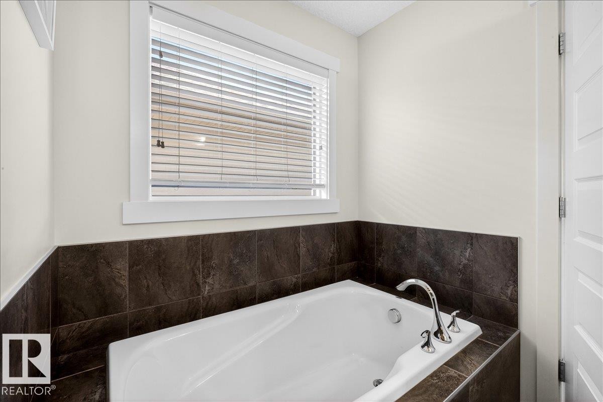Spa-inspired primary bath featuring a deep soaking tub with chrome fixtures, dark tile wainscoting, crisp white upper walls, and a horizontal blinds window for natural light and privacy - 3936 Claxton Loop Sw, Edmonton, AB - Indoor Photo Showing Bathroom