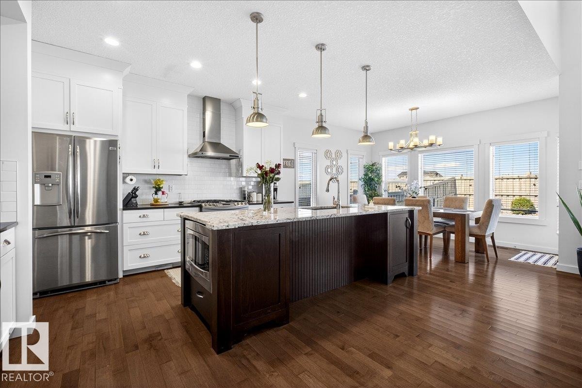 Gourmet kitchen featuring stainless steel French-door refrigerator and gas range with hood ventilation, white cabinetry with brushed nickel hardware, quartz countertops, and a substantial dark wood-finish island with pendant lighting - 3936 Claxton Loop Sw, Edmonton, AB - Indoor Photo Showing Kitchen With Stainless Steel Kitchen With Upgraded Kitchen