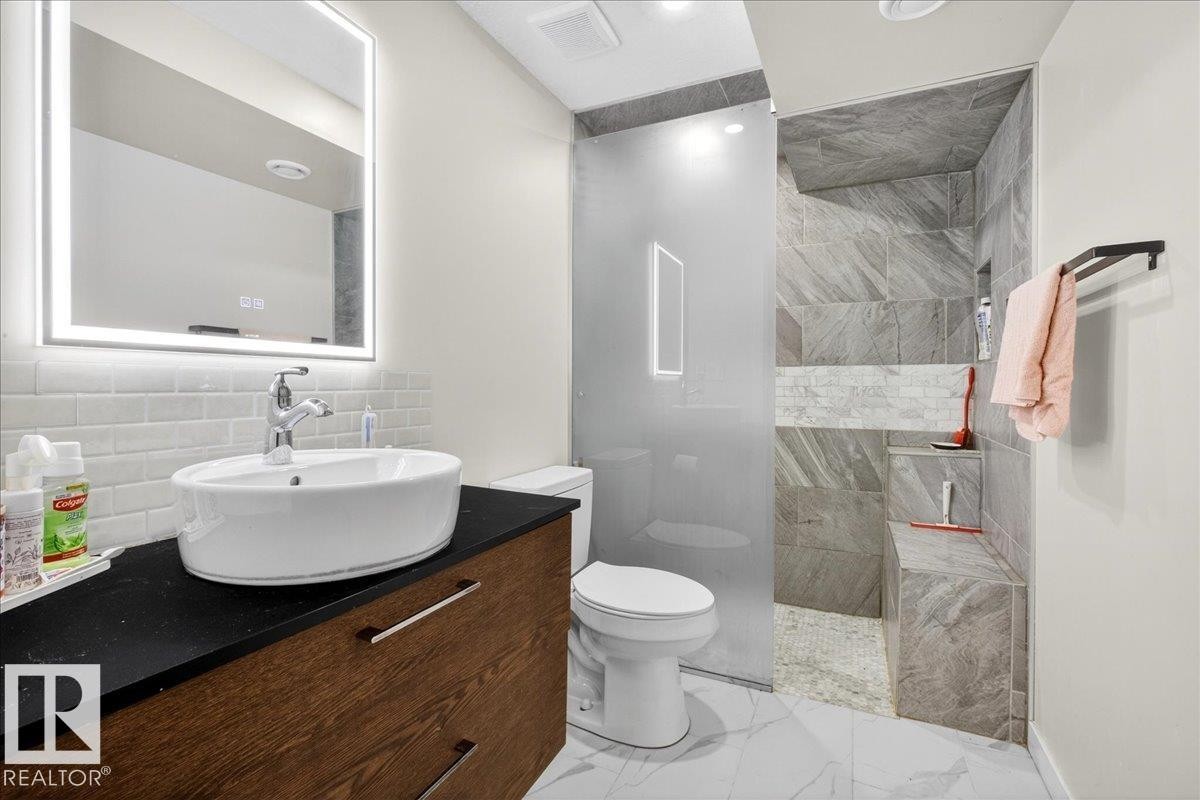 Backlit vanity mirror, vessel sink, wood-finish vanity, subway tile backsplash, and a tiled shower with a built-in bench - 3936 Claxton Loop Sw, Edmonton, AB - Indoor Photo Showing Bathroom