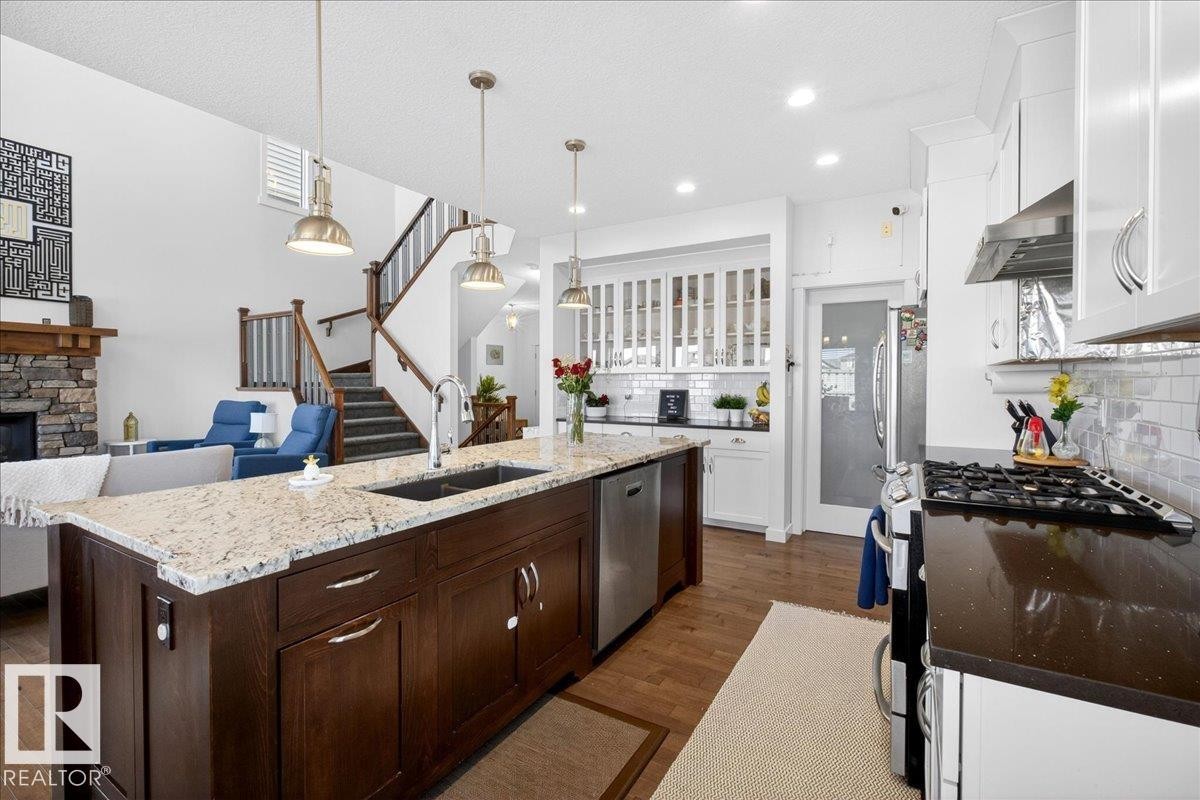 Chef's kitchen featuring a substantial dark wood island with light stone countertops, professional-grade gas range with stainless steel hood, white cabinetry with glass-front uppers, recessed lighting, and brass pendant fixtures - 3936 Claxton Loop Sw, Edmonton, AB - Indoor Photo Showing Kitchen With Double Sink With Upgraded Kitchen