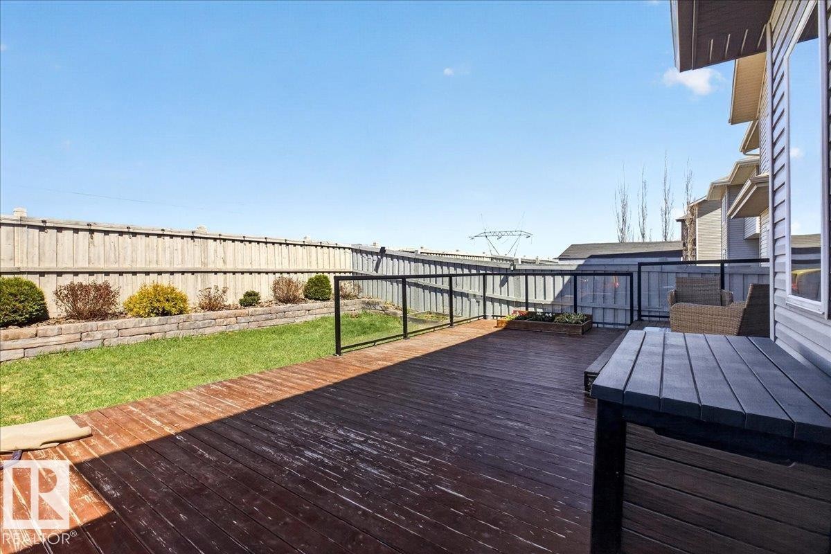Expansive wooden deck with glass panel railing, featuring a manicured lawn and stone retaining wall - 3936 Claxton Loop Sw, Edmonton, AB - Outdoor With Exterior