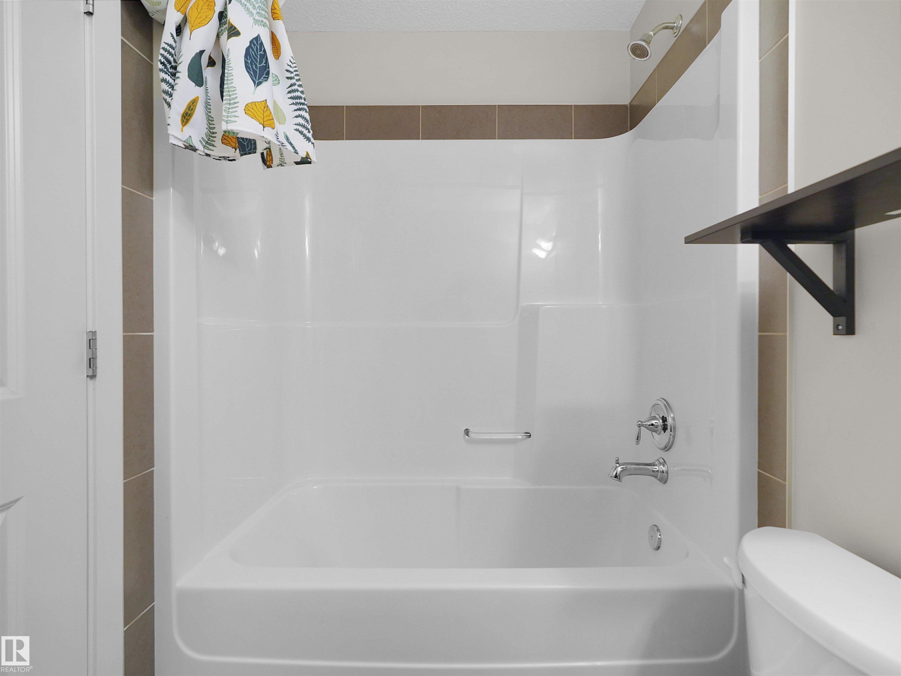 White bathtub with integrated shower, chrome fixtures, and neutral-toned wall tiles - #89 603 Watt Bv Sw, Edmonton, AB - Indoor Photo Showing Bathroom