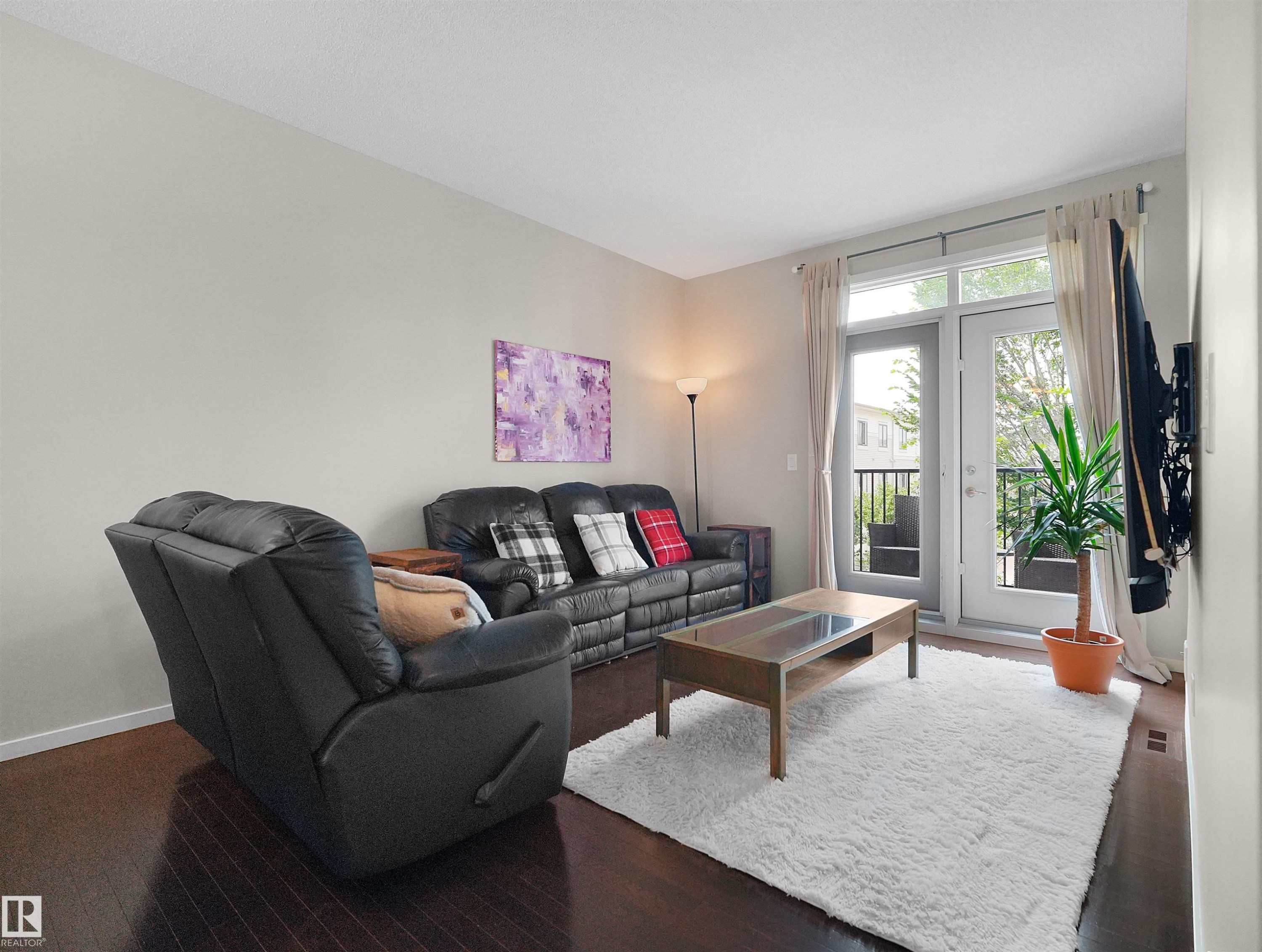 Dark wood-finish flooring, light neutral wall tones, and double French doors opening to an exterior balcony with black iron railings - #89 603 Watt Bv Sw, Edmonton, AB - Indoor Photo Showing Living Room