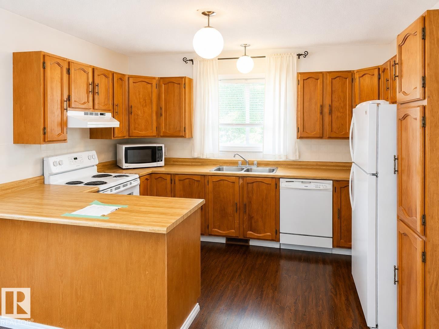Kitchen featuring wood-finish cabinetry, a built-in double basin sink, white appliances, and wood-finish flooring - 802 16 Avenue, Cold Lake, AB - Indoor Photo Showing Kitchen With Double Sink