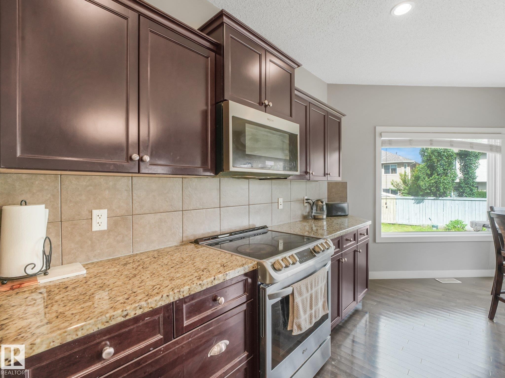 Kitchen featuring dark wood cabinetry, light-toned tiled backsplash, speckled stone countertops, and stainless steel appliances - 1478 37B Avenue Nw, Edmonton, AB - Indoor Photo Showing Kitchen With Upgraded Kitchen