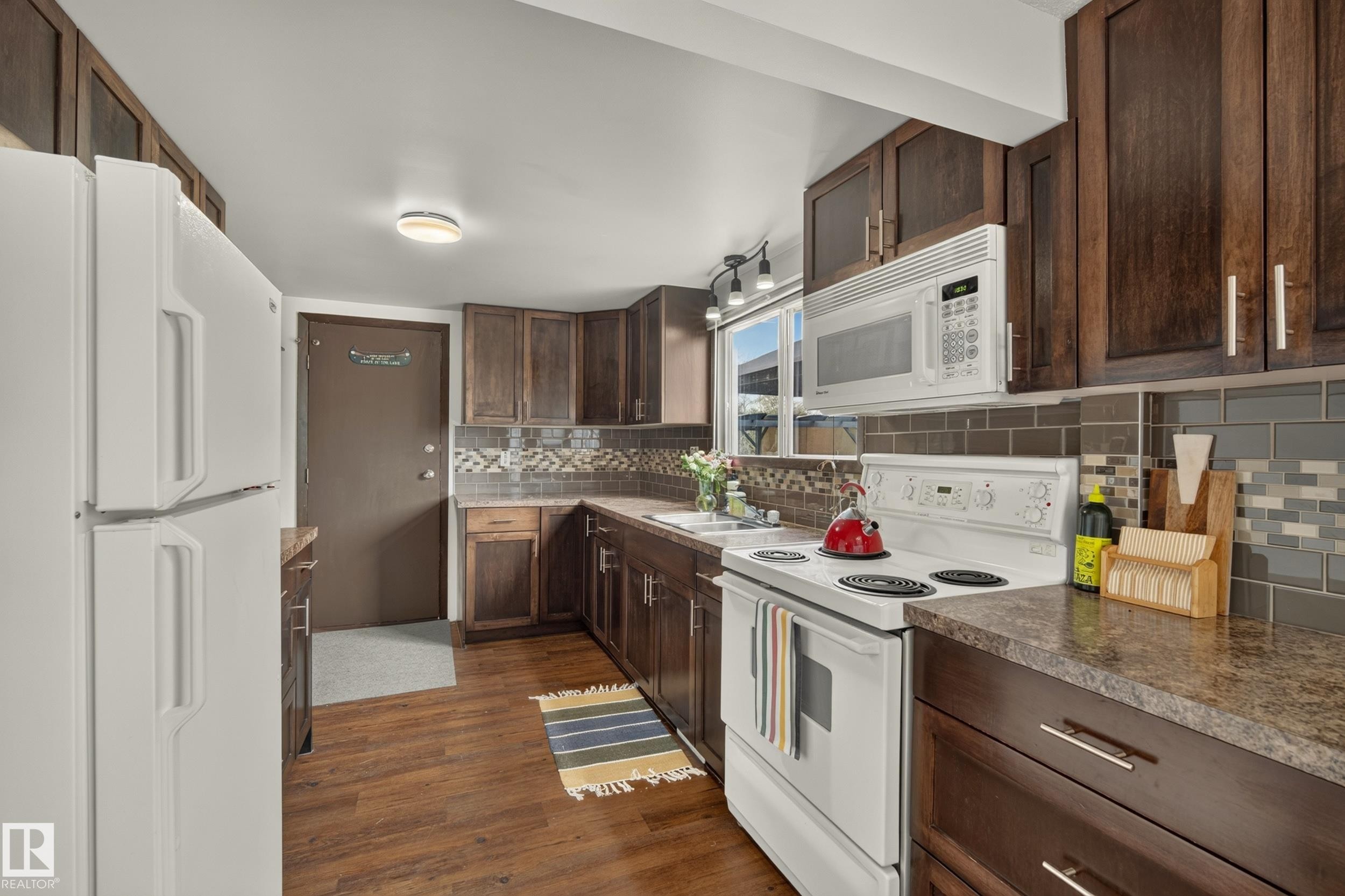 Wood-finish flooring, dark wood cabinetry, composite countertops, white appliances, and a tiled backsplash - 264 Beach Avenue, Rural Barrhead County, AB - Indoor Photo Showing Kitchen