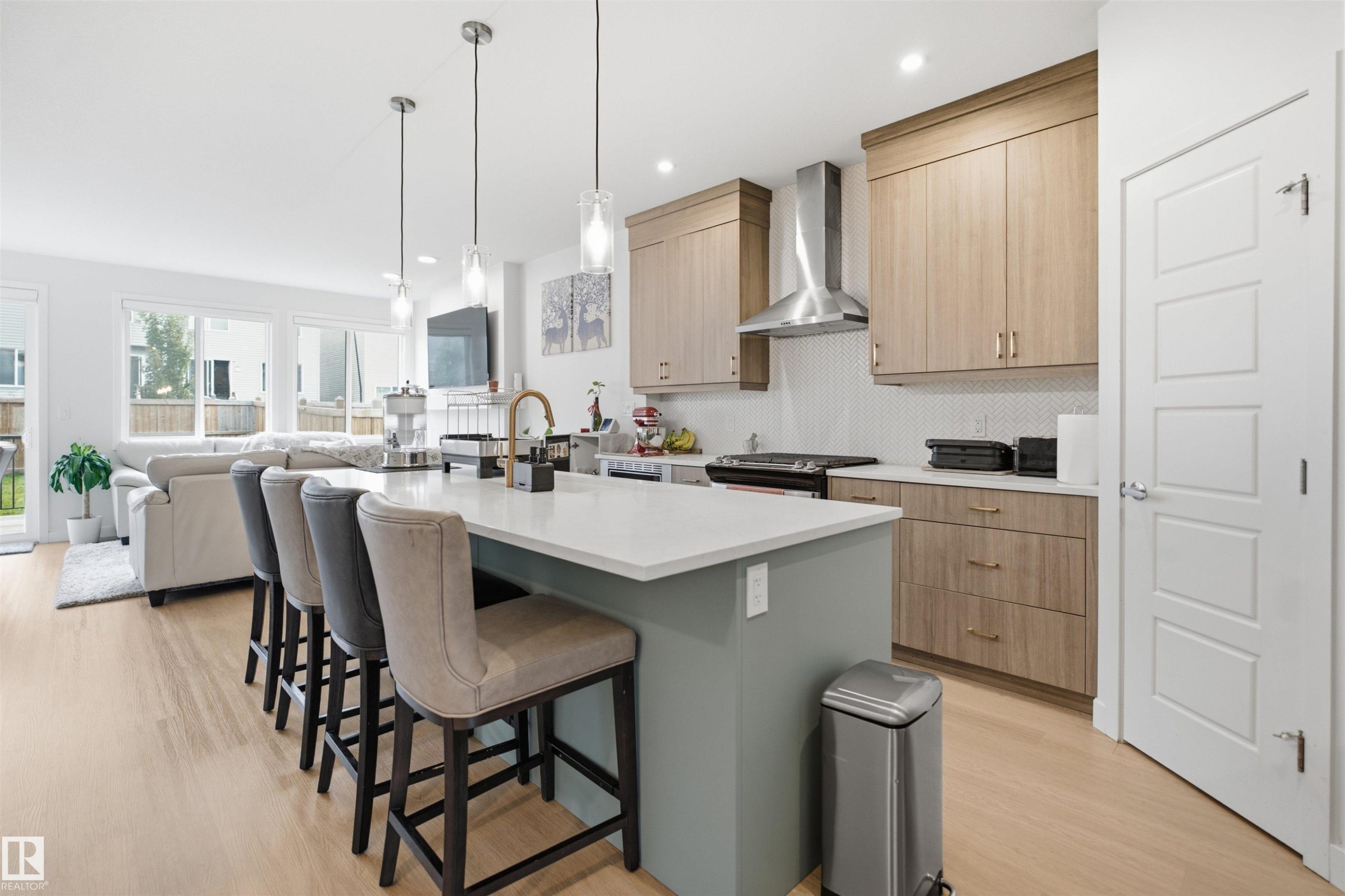 Kitchen island with a white countertop and pendant lighting, wood-finish flooring, light wood-finish cabinetry, stainless steel range hood, and white panel doors - 3877 Chrustawka Place Sw, Edmonton, AB - Indoor