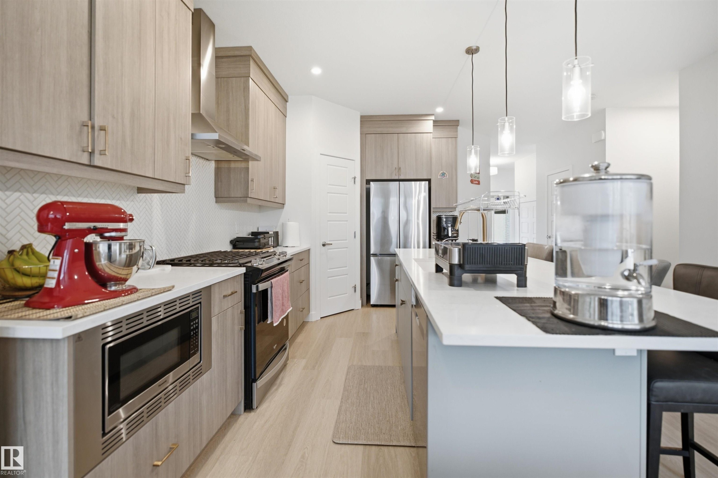 Modern kitchen featuring wood-finish cabinetry, stainless steel appliances, a white herringbone backsplash, and a large island with pendant lighting - 3877 Chrustawka Place Sw, Edmonton, AB - Indoor Photo Showing Kitchen With Upgraded Kitchen