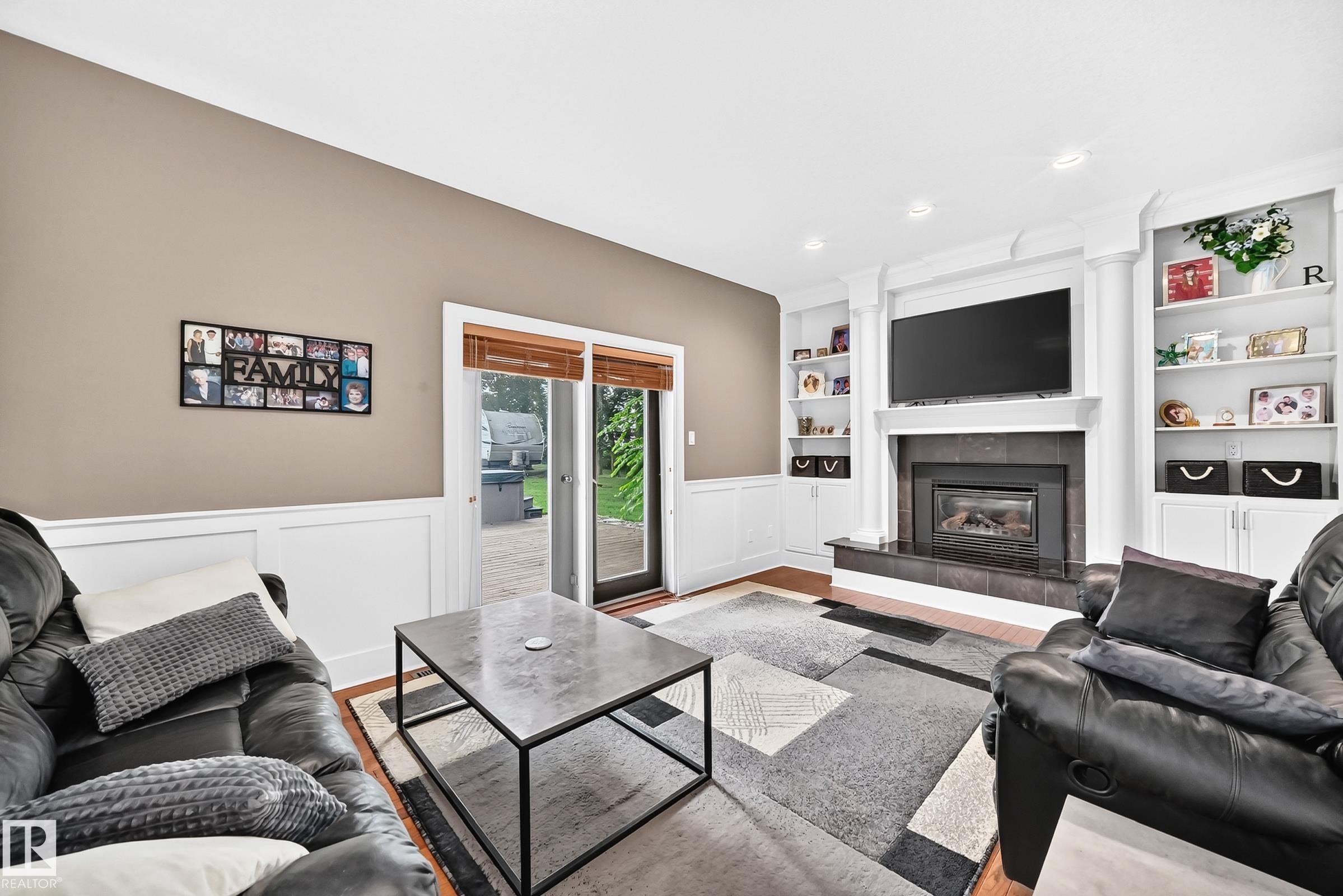 Fireplace with dark tile surround, built-in shelving, white wainscoting, sliding glass door, and wood-finish flooring - #38 24512 Sturgeon Rd, Rural Sturgeon County, AB