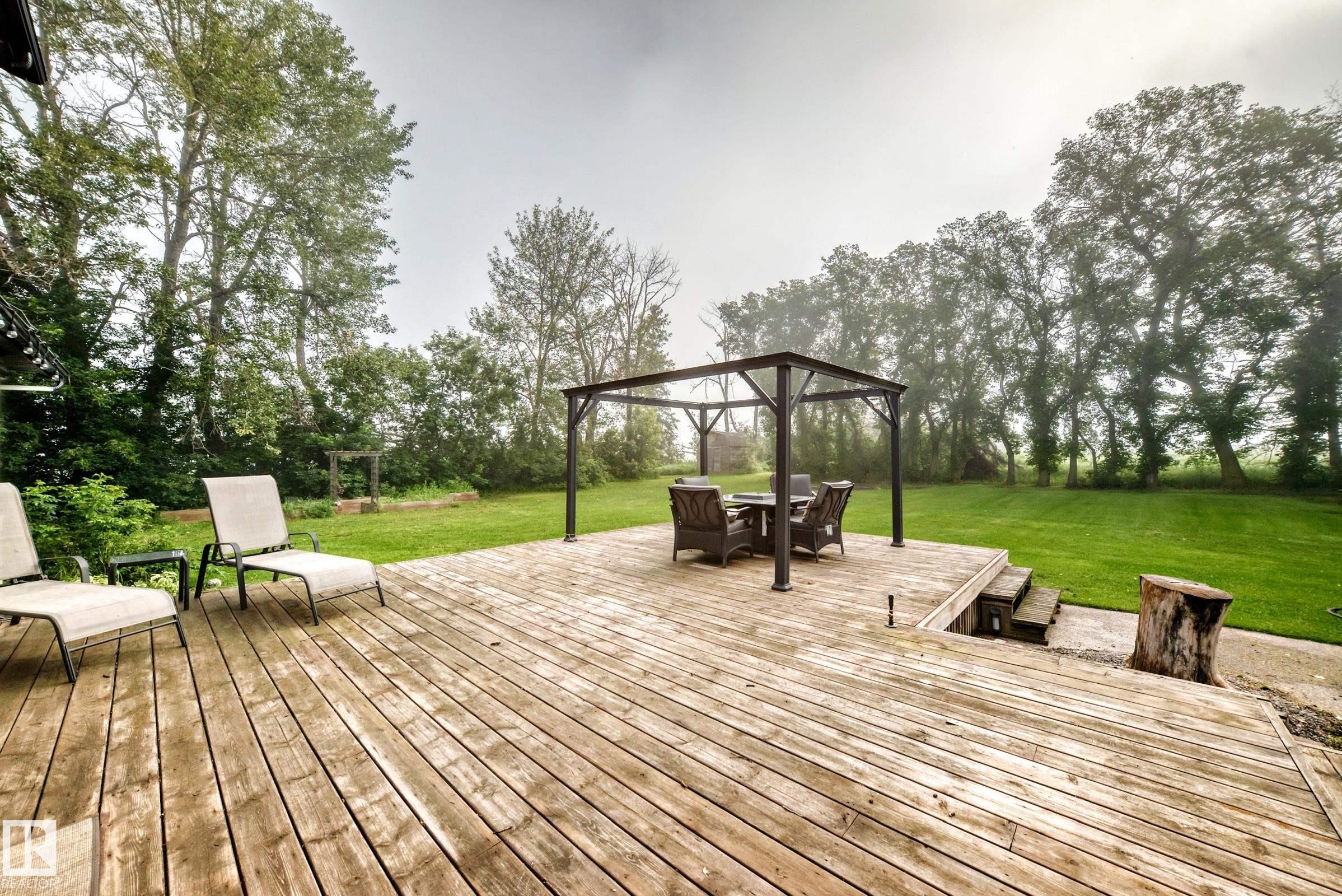 Expansive wood-finish deck with a metal gazebo and integrated steps leading to a vast green lawn, bordered by mature trees - #38 24512 Sturgeon Rd, Rural Sturgeon County, AB