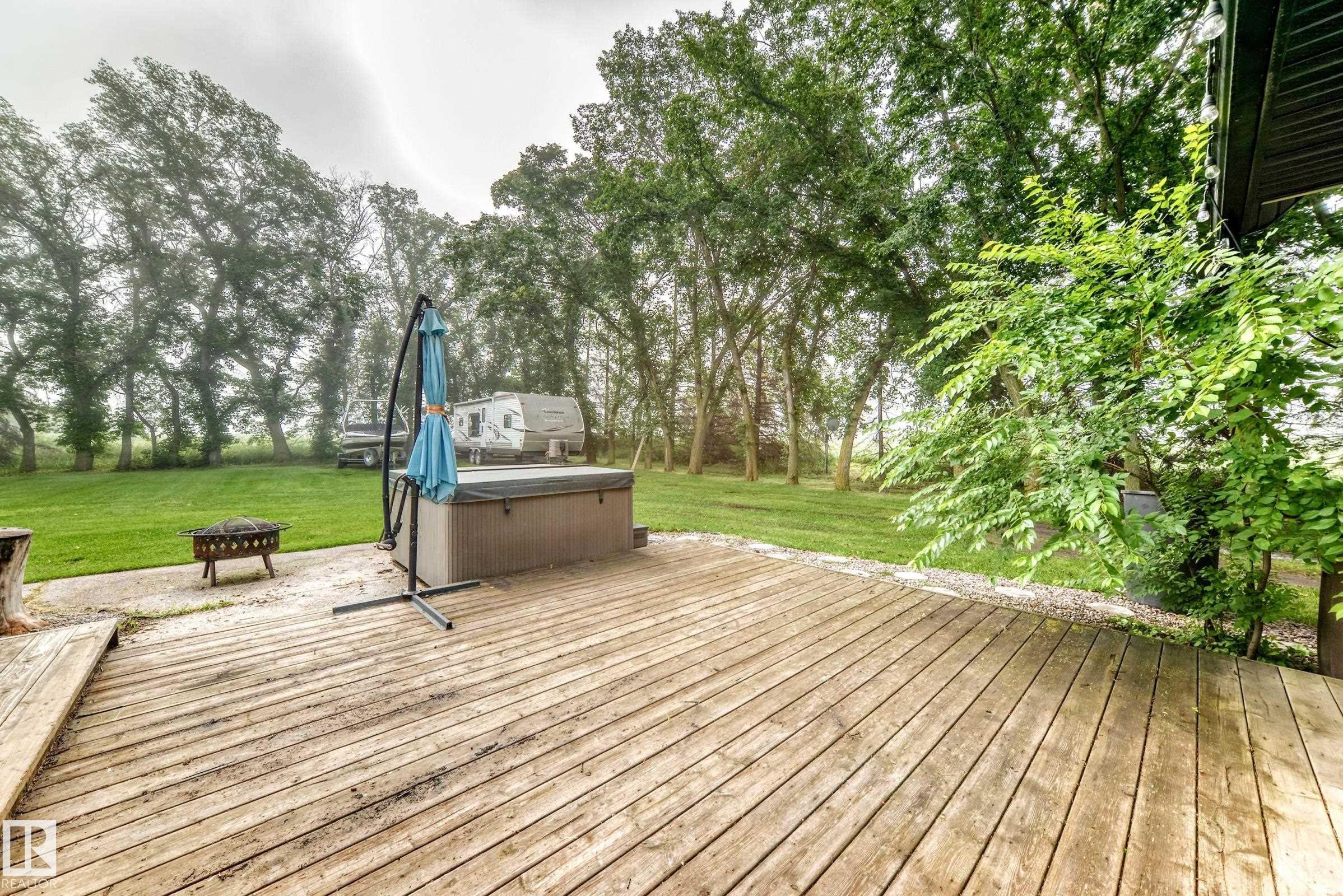 Expansive wood deck with a built-in spa, gravel hardscaping, and a lush green lawn surrounded by mature trees - #38 24512 Sturgeon Rd, Rural Sturgeon County, AB