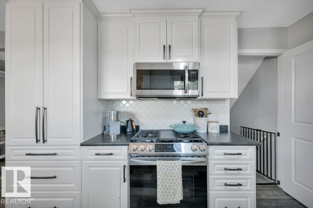 White cabinetry with dark pulls, stainless steel appliances, dark countertop, and decorative tile backsplash - #138 5231 51 St, Bon Accord, AB - Indoor Photo Showing Kitchen