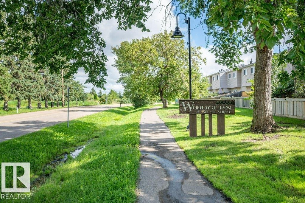 Paved pathway bordered by manicured lawn and mature trees, featuring a community sign and street lamp - #138 5231 51 St, Bon Accord, AB - Outdoor