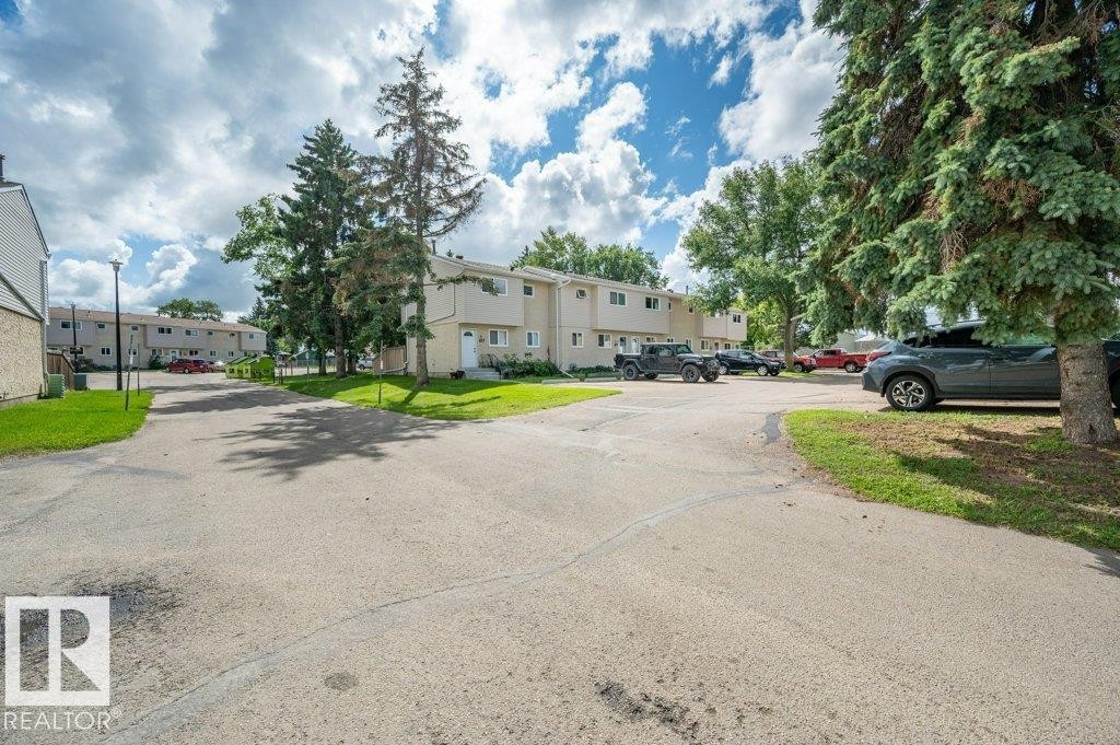 Paved parking area surrounded by mature trees and grass, with multi-unit structures in the background - #138 5231 51 St, Bon Accord, AB - Outdoor