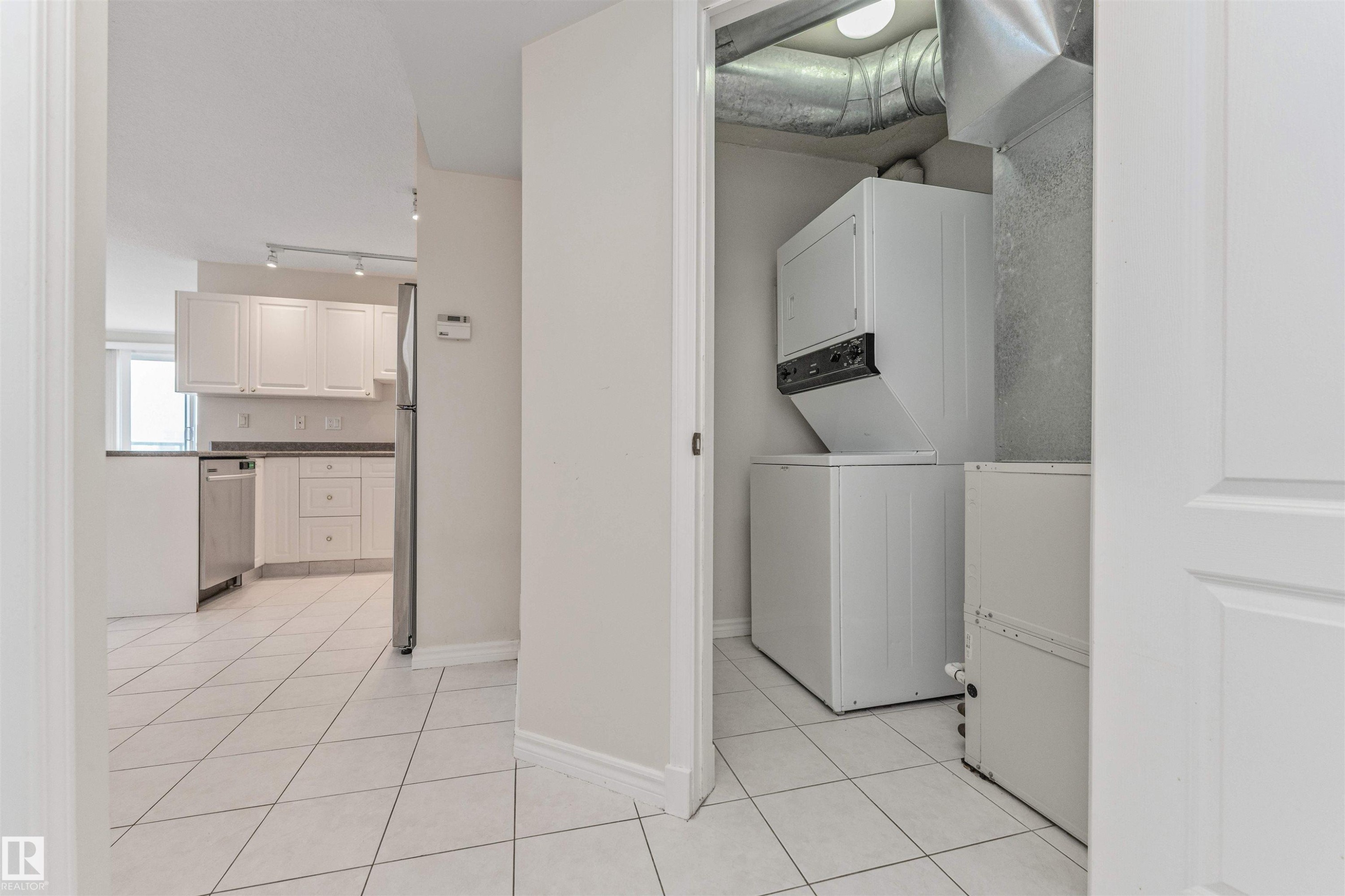 White tile flooring throughout, with a stacked washer and dryer unit, and a kitchen featuring white cabinetry, stainless steel appliances, and dark countertops - #1101 10909 103 Av Nw, Edmonton, AB - Indoor Photo Showing Laundry Room