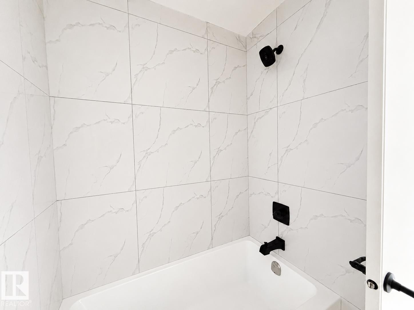 Bathtub featuring a white finish, large format wall tiling with subtle veining, matte black showerhead, and matte black bath fixtures - 13118 186 Avenue Nw, Edmonton, AB - Indoor Photo Showing Bathroom