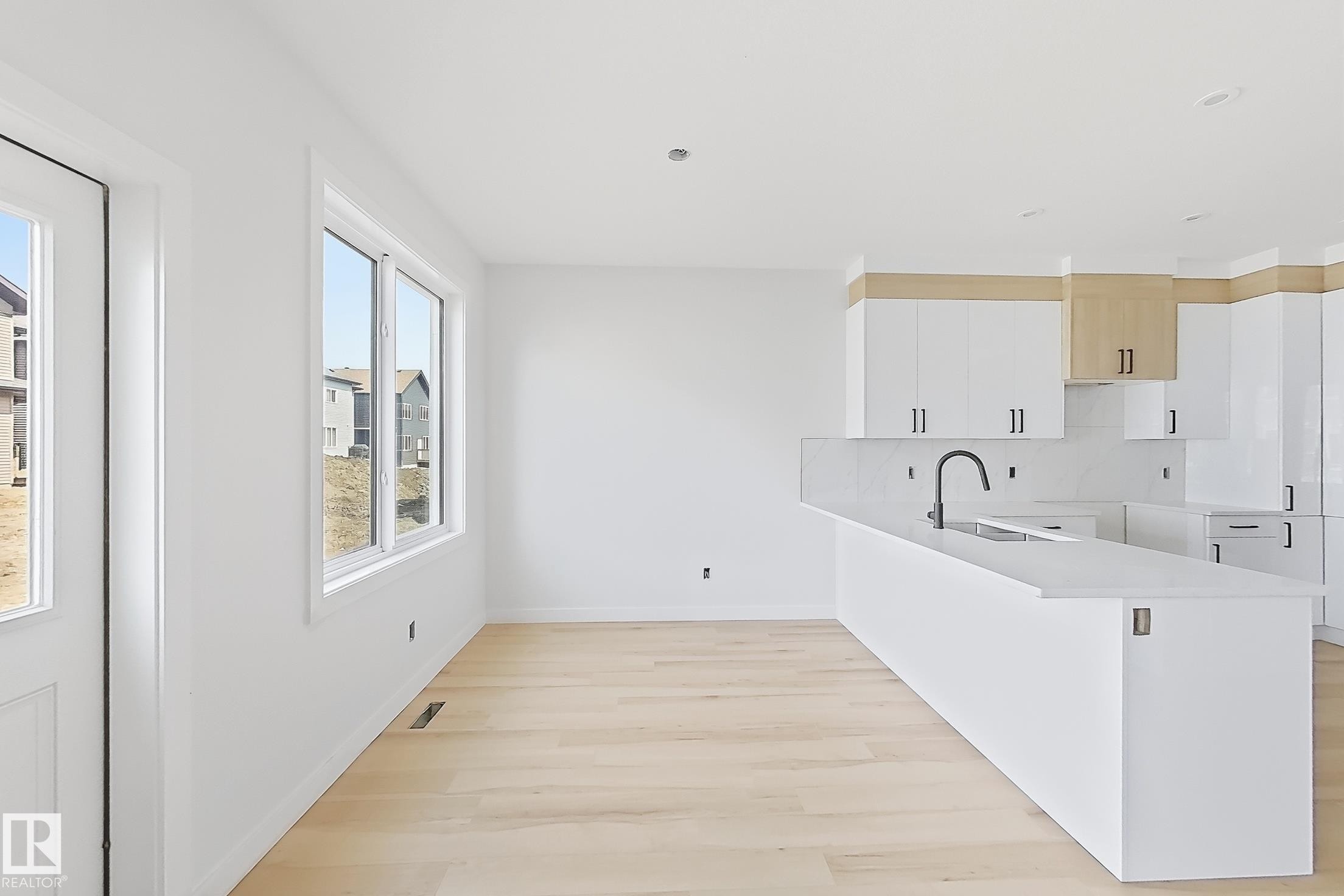 Modern kitchen featuring a large island with sink and matte black faucet, light wood-finish flooring, white cabinetry with contrasting wood-finish accents, and multiple windows - 13118 186 Avenue Nw, Edmonton, AB - Indoor