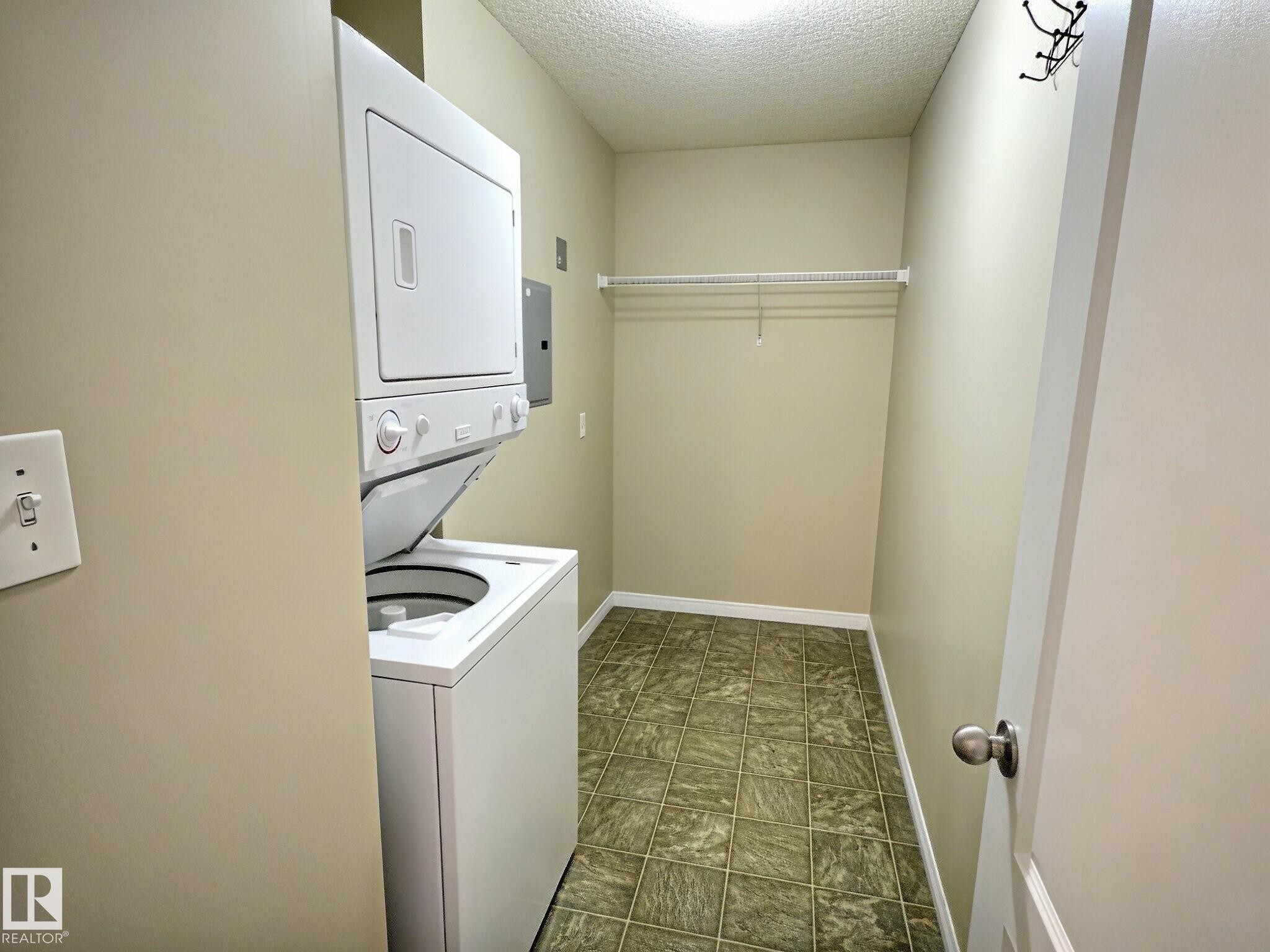 Laundry area with a stacked washer and dryer, patterned floor tiling, and a wire shelving unit - #111 5350 199 St Nw, Edmonton, AB - Indoor Photo Showing Laundry Room