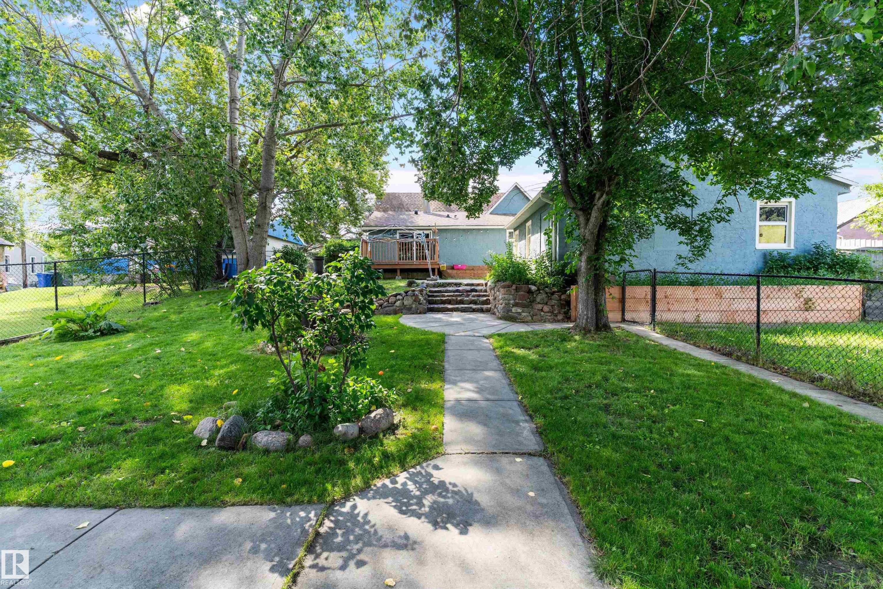 Manicured lawn with mature trees, paved pathway, stone retaining wall, and wood deck - 5404 45 Avenue, Wetaskiwin, AB - Outdoor
