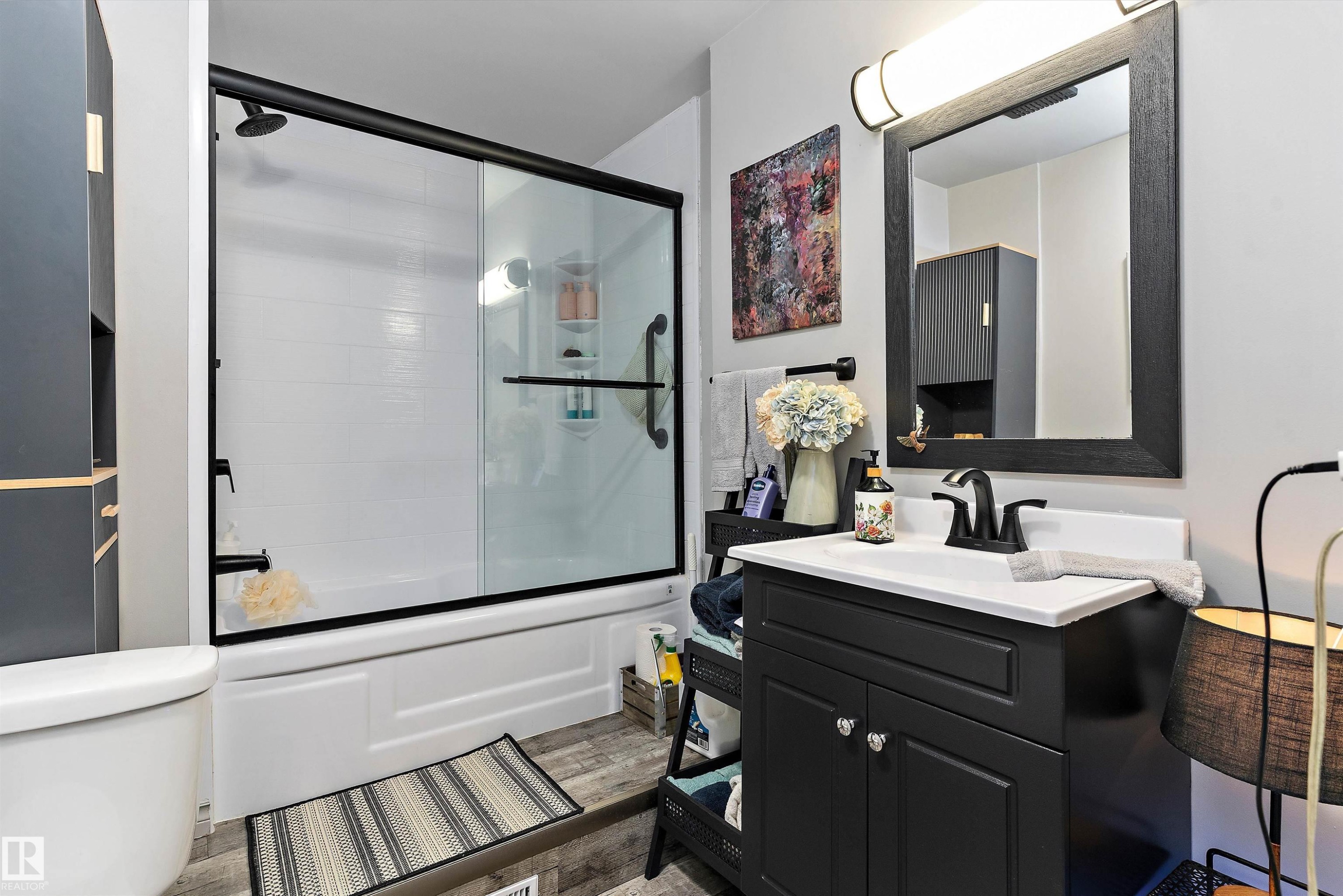 White vanity with a solid surface countertop and matte black faucet, framed mirror with integrated lighting, built-in shower over tub with a glass enclosure, and wood-finish flooring - 5404 45 Avenue, Wetaskiwin, AB - Indoor Photo Showing Bathroom