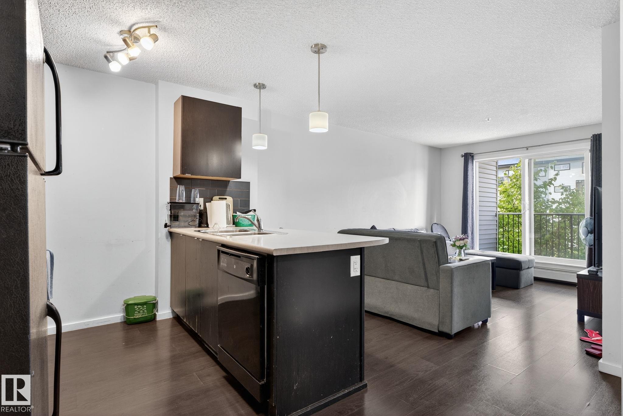Open-concept living area featuring dark wood-finish flooring, a kitchen island with a light-toned countertop, pendant lighting fixtures, and a glass sliding door leading to an outdoor balcony - 3353 16A Avenue Nw, Edmonton, AB - Indoor