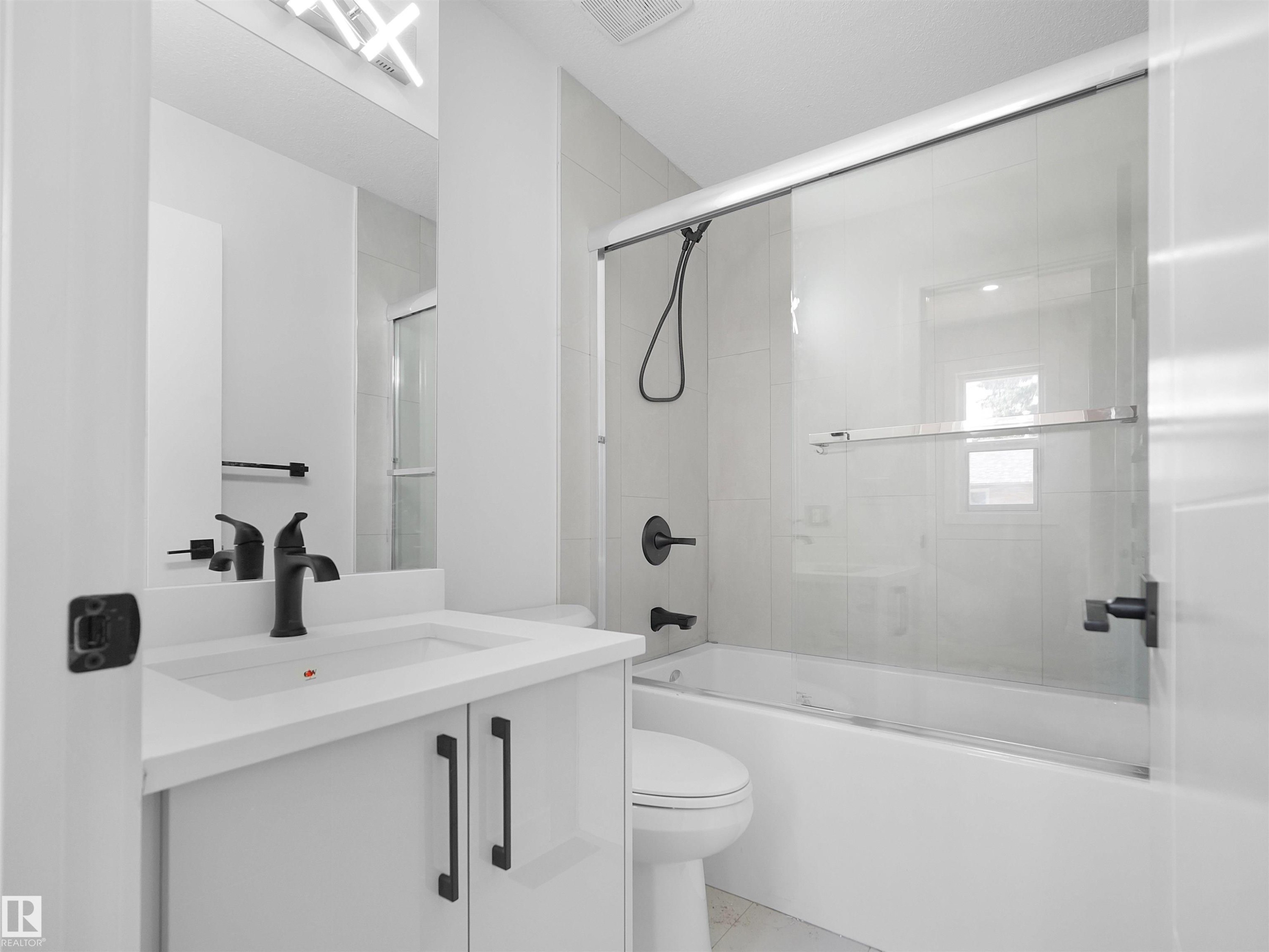 Bathroom featuring a white vanity with integrated sink, matte black fixtures, an illuminated mirror, a toilet, and a bathtub with a clear glass sliding door - 126 Woodstock Nw, Edmonton, AB - Indoor Photo Showing Bathroom