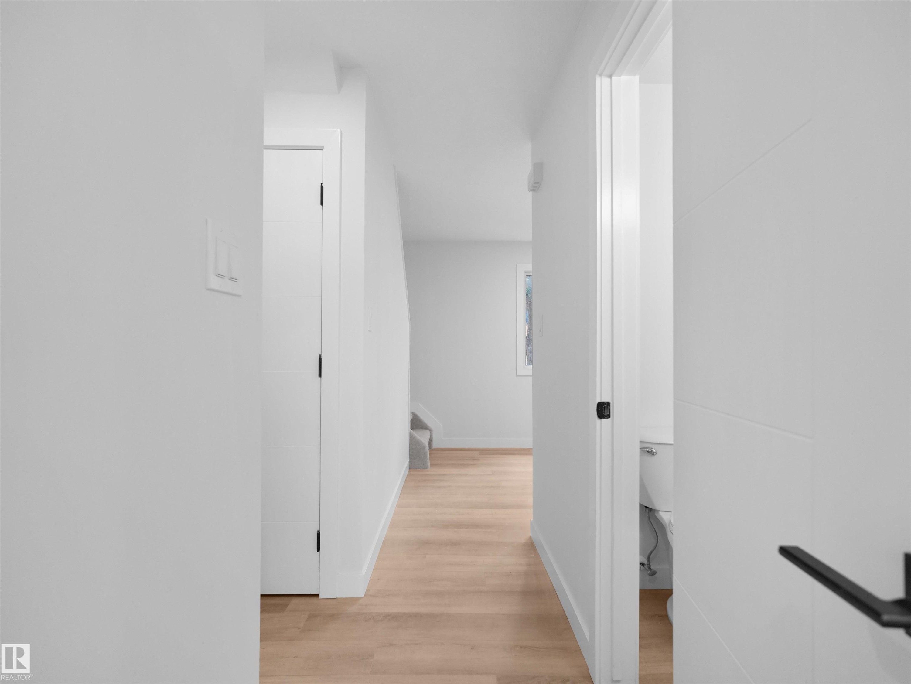 Light wood-finish flooring, white walls, modern interior doors with matte black hardware, and a visible toilet in an adjacent room - 126 Woodstock Nw, Edmonton, AB - Indoor Photo Showing Other Room