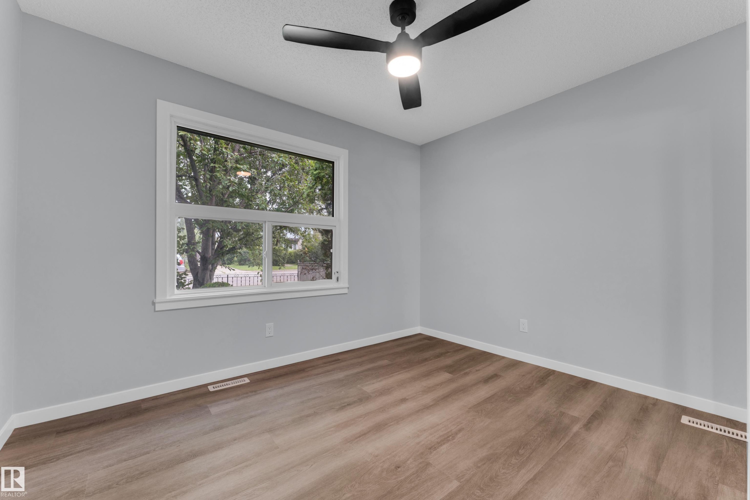 Bright room featuring a large window with white trim, wood-finish flooring, light gray walls, and a contemporary black ceiling fan with integrated lighting - 9416 153 Street Nw, Edmonton, AB - Indoor Photo Showing Other Room