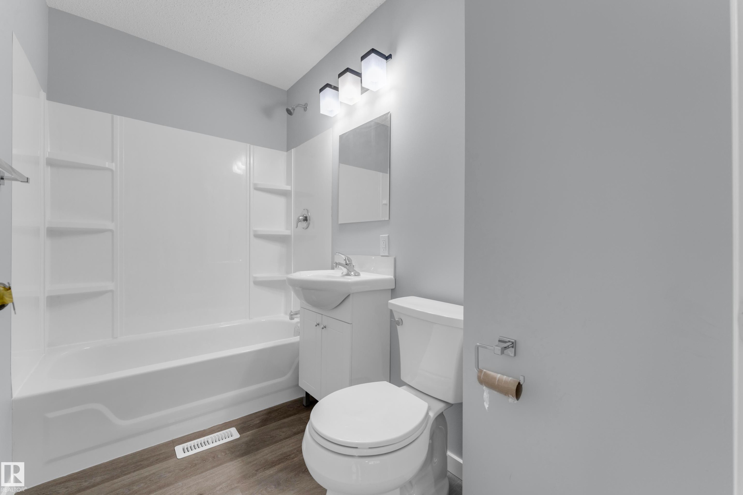 Bathroom featuring a white tub/shower combination with integrated shelving, a compact vanity with a white sink, a mirror, and wood-finish flooring - 9416 153 Street Nw, Edmonton, AB - Indoor Photo Showing Bathroom