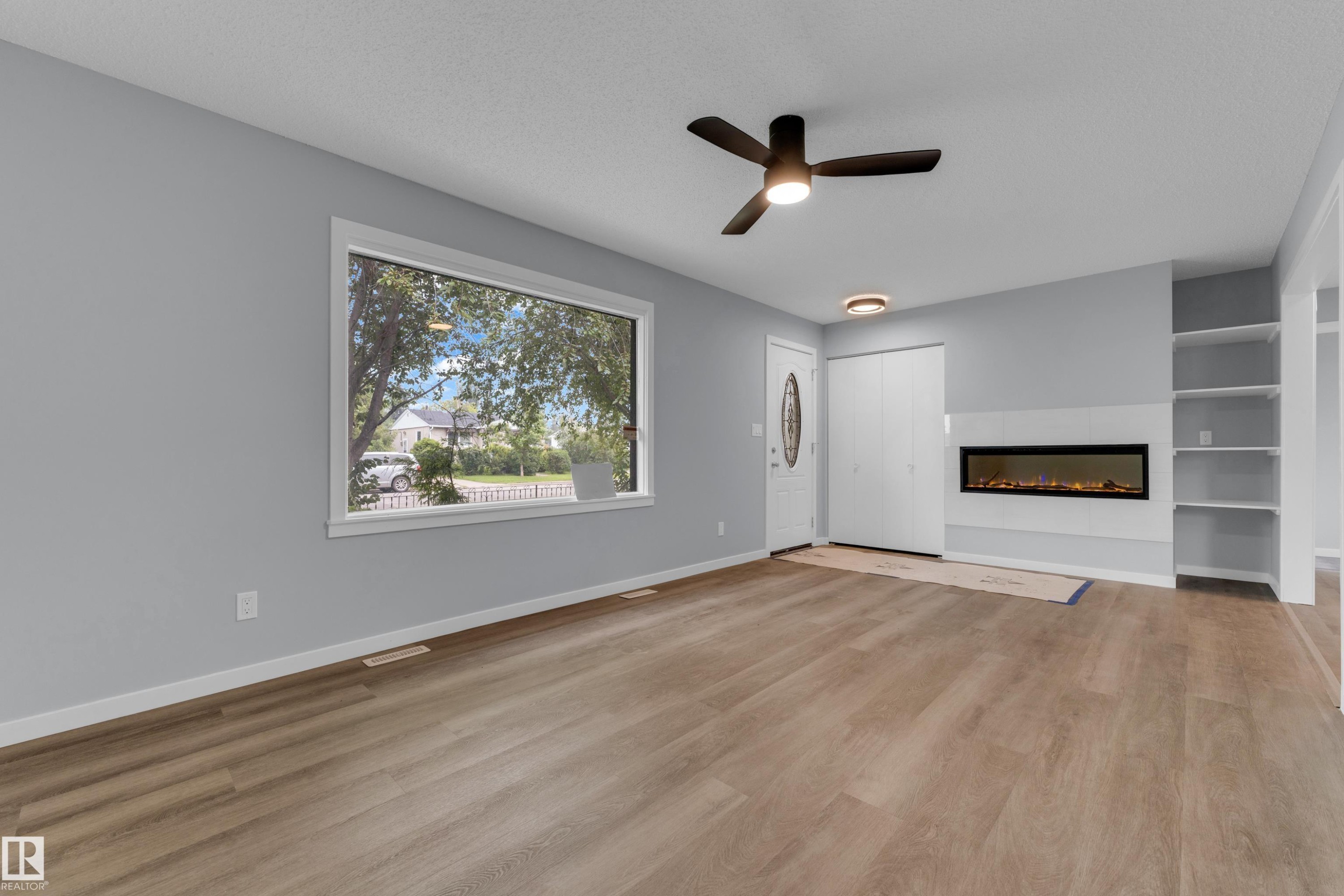 Spacious living area featuring wood-finish flooring, a contemporary ceiling fan with integrated lighting, a large picture window, an electric fireplace with a white surround, and built-in shelving - 9416 153 Street Nw, Edmonton, AB - Indoor With Fireplace