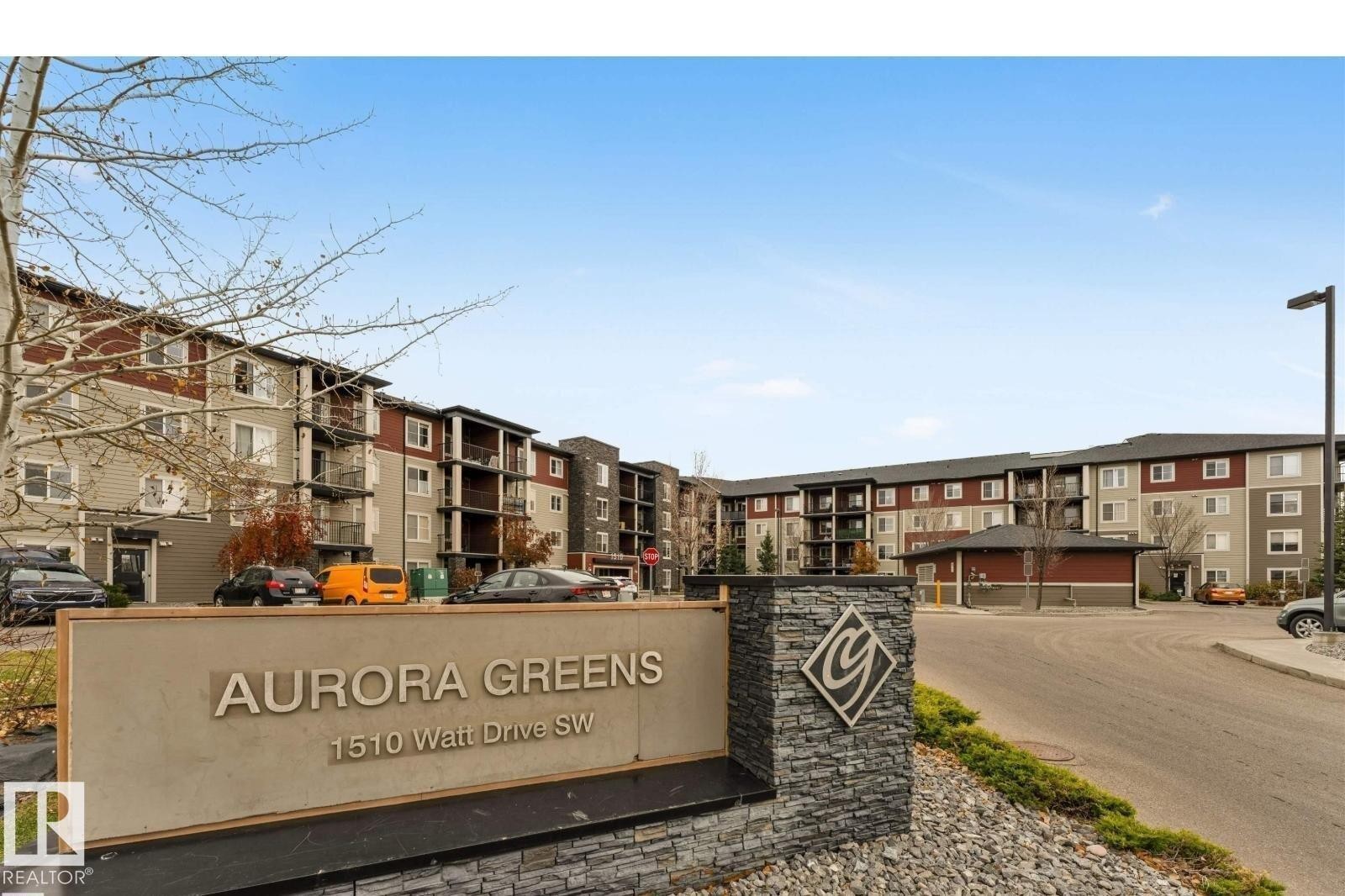 Aurora Greens residential community featuring multi-story apartment buildings with brick and stone exterior finishes, organized street-facing layout, and professional landscaping with mature trees and shrub plantings along the entrance signage - #118 1510 Watt Dr Sw, Edmonton, AB - Outdoor With Facade