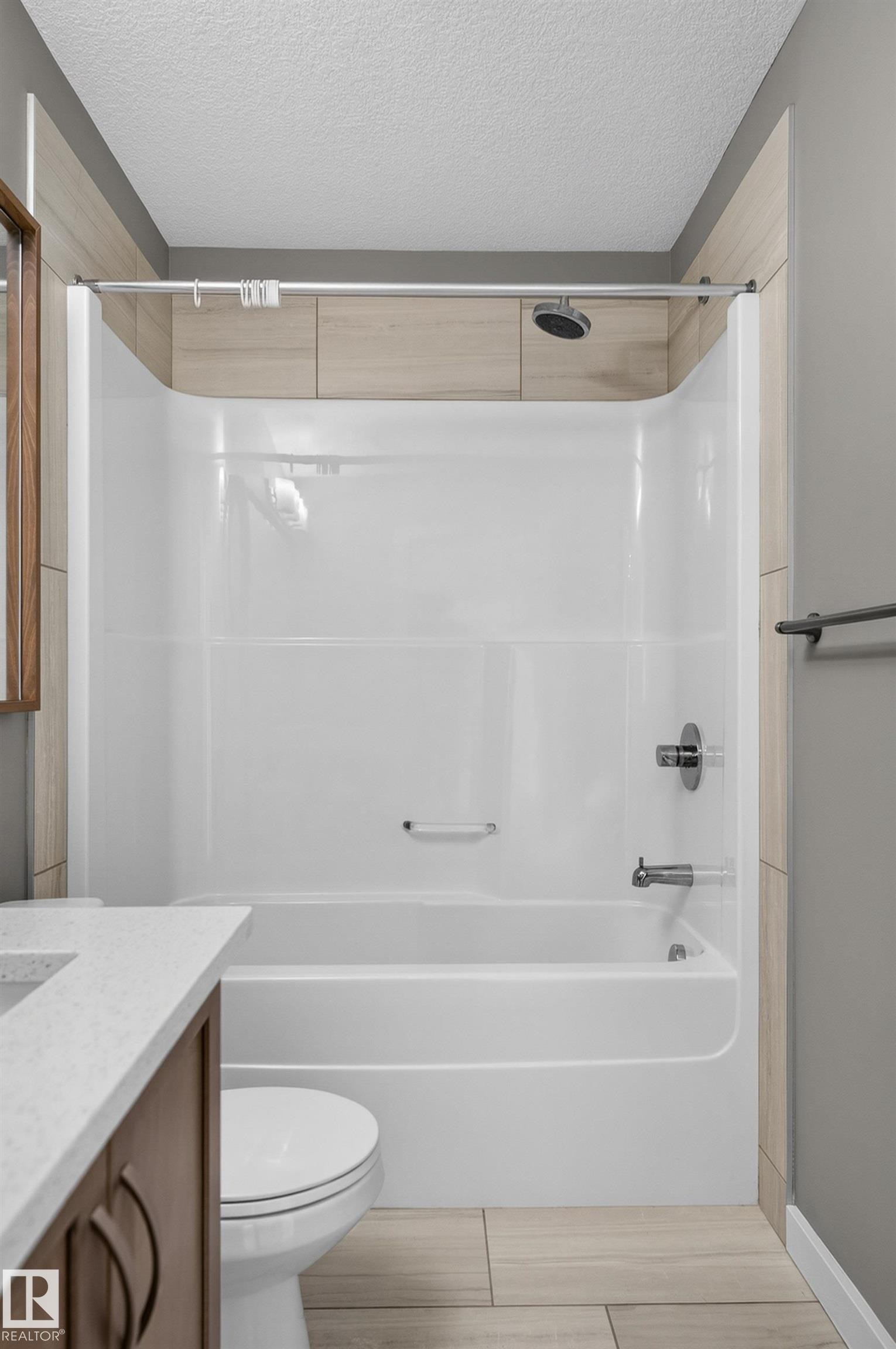 Bathtub with shower fixture, wood-finish wall tile, and light-toned floor tiles - #131 5504 Schonsee Dr Nw, Edmonton, AB - Indoor Photo Showing Bathroom