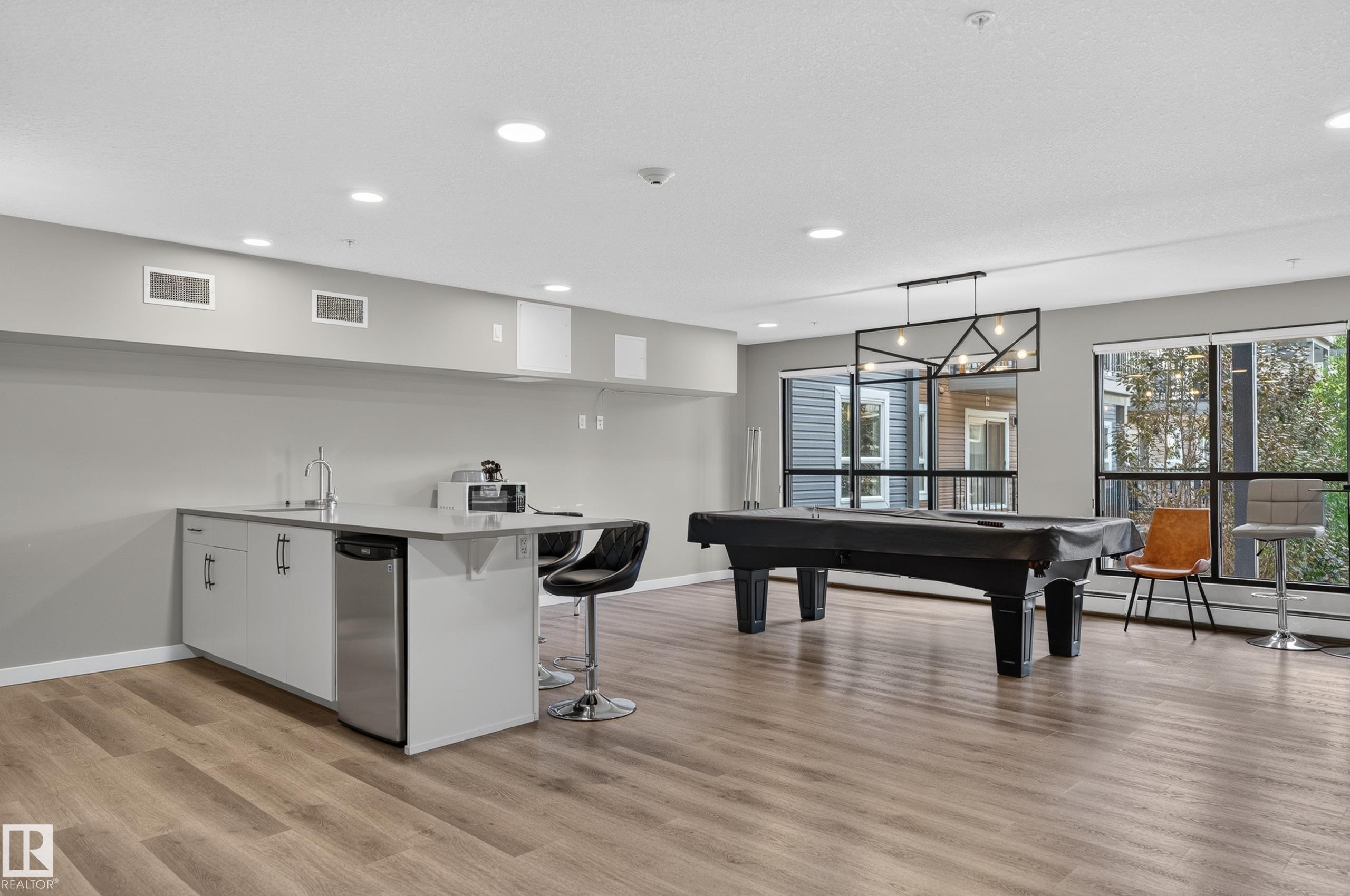 Expansive common area featuring wood-finish flooring, a built-in wet bar with stainless steel appliances, large windows, and recessed lighting - #131 5504 Schonsee Dr Nw, Edmonton, AB - Indoor Photo Showing Other Room