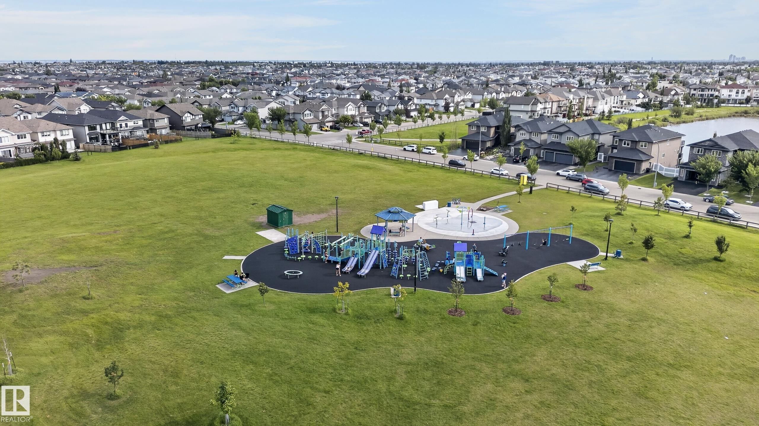 Expansive green space featuring a playground with multiple structures, a splash pad area, and a paved pathway - #131 5504 Schonsee Dr Nw, Edmonton, AB - Outdoor With View