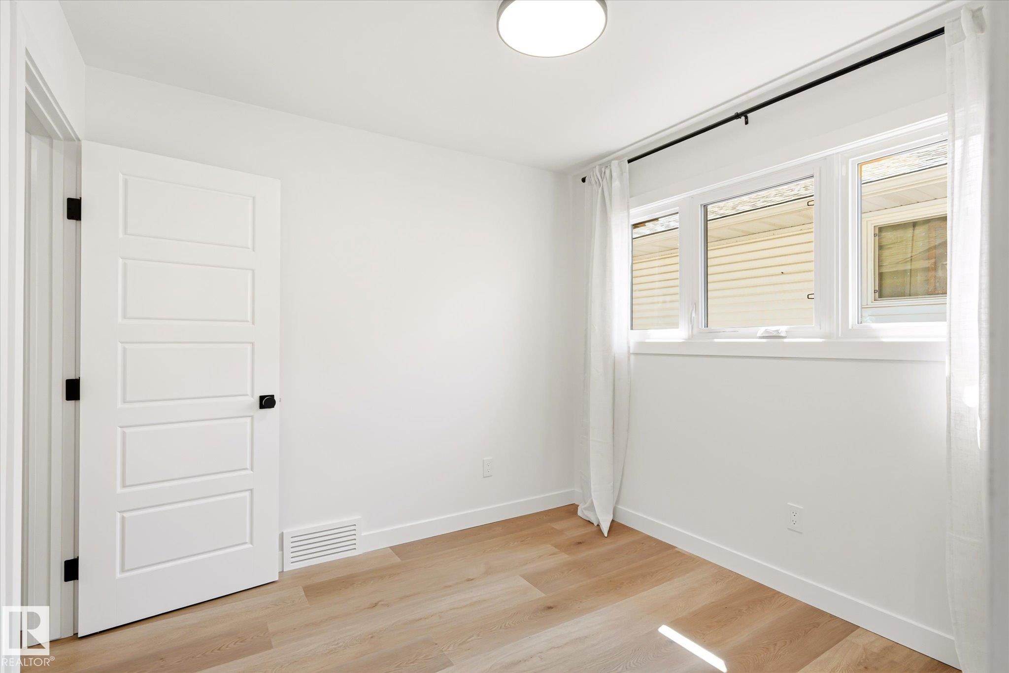 Room featuring wood-finish flooring, a white five-panel door with black hardware, a three-pane window with white trim, a black curtain rod, and a flush-mount ceiling light fixture - 4708 104A Avenue Nw, Edmonton, AB - Indoor Photo Showing Other Room
