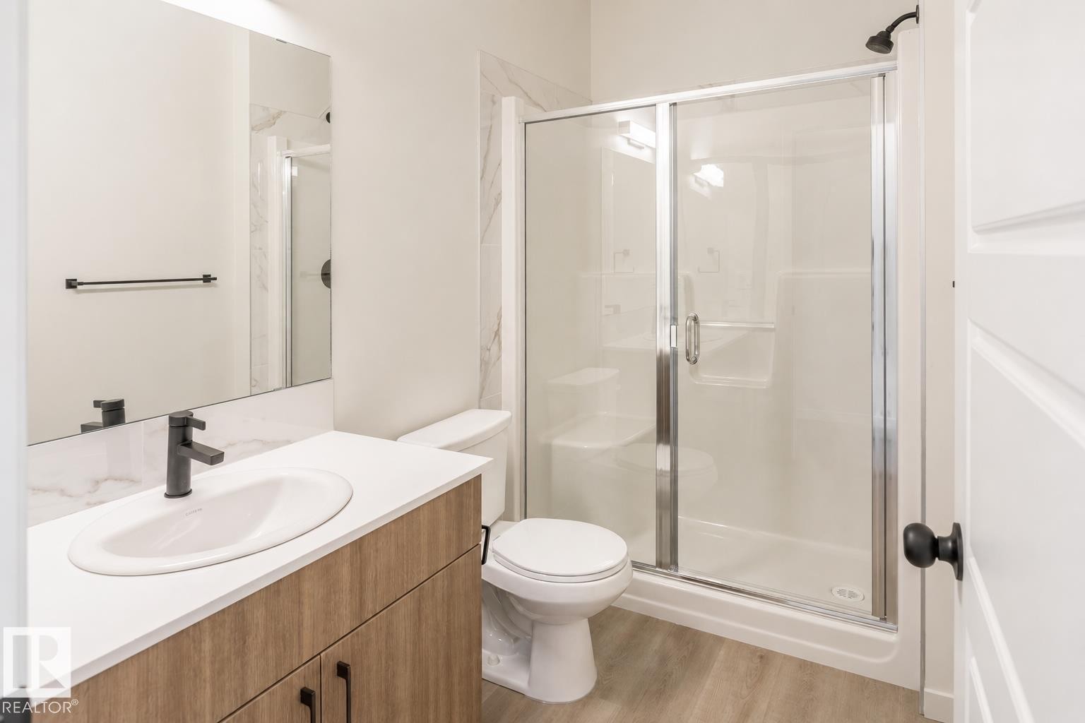 Wood-finish vanity with an integrated sink and matte black faucet, glass-enclosed shower with a built-in bench, and light wood-finish flooring - 219 157 Avenue Ne, Edmonton, AB - Indoor Photo Showing Bathroom