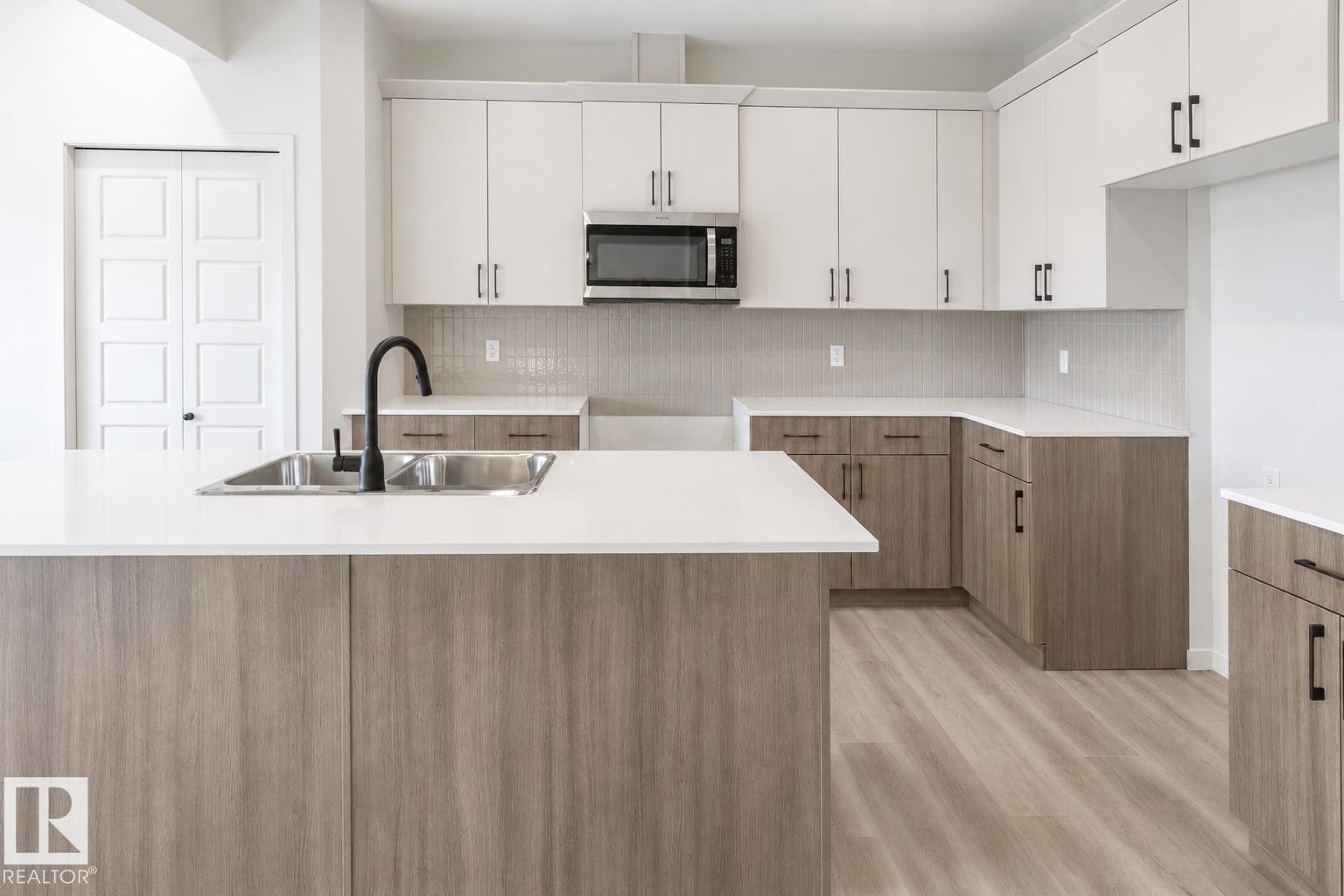 Spacious kitchen featuring an island with an undermount double-basin sink, a matte black faucet, light wood-finish lower cabinetry, white upper cabinetry, and light-toned countertops - 219 157 Avenue Ne, Edmonton, AB - Indoor Photo Showing Kitchen With Double Sink