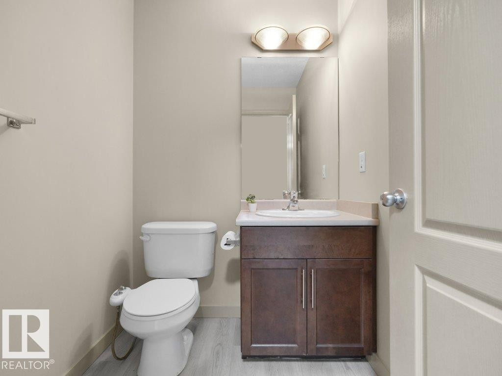 Wood-finish flooring with a white toilet, dark wood-finish vanity, integrated sink, and wall-mounted mirror - 3010 17A Ave Nw, Edmonton, AB - Indoor Photo Showing Bathroom