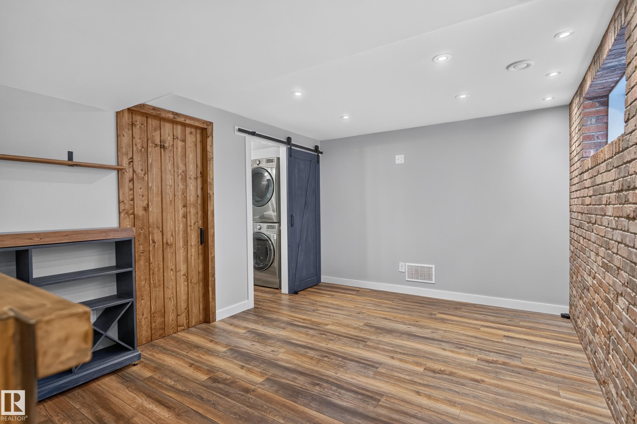 Wood-finish flooring, exposed brick wall, recessed ceiling lighting, and a barn door leading to a laundry area with stacked appliances - 6008 South Terwillegar Boulevard Nw, Edmonton, AB - Indoor Photo Showing Other Room