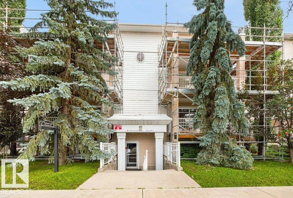 Building entrance with prominent signage, framed by mature evergreen trees - #201 10528 77 Av Nw, Edmonton, AB - Outdoor