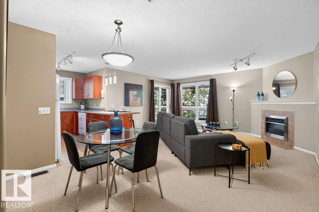 Open-concept living area featuring a fireplace, multiple windows, kitchen with wood-finish cabinetry, and a dedicated dining space with pendant lighting - #201 10528 77 Av Nw, Edmonton, AB - Indoor With Fireplace